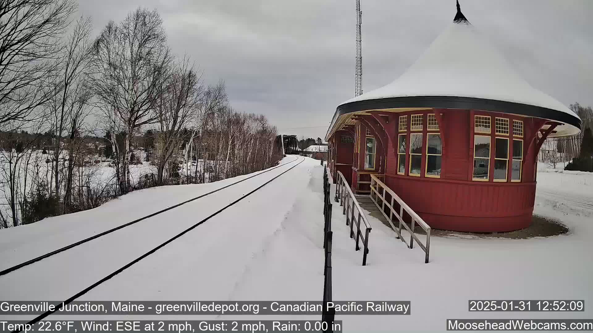 A snow-covered train platform with a red, round train station sits beside a set of train tracks leading towards a bridge under a cloudy sky.