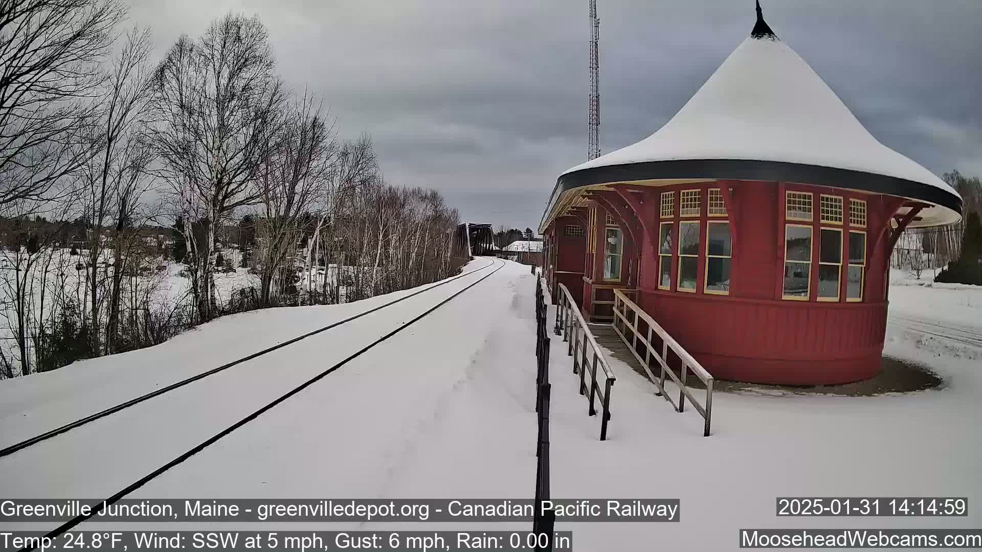 A snow-covered railway track leads to a red, round train station under a cloudy sky.