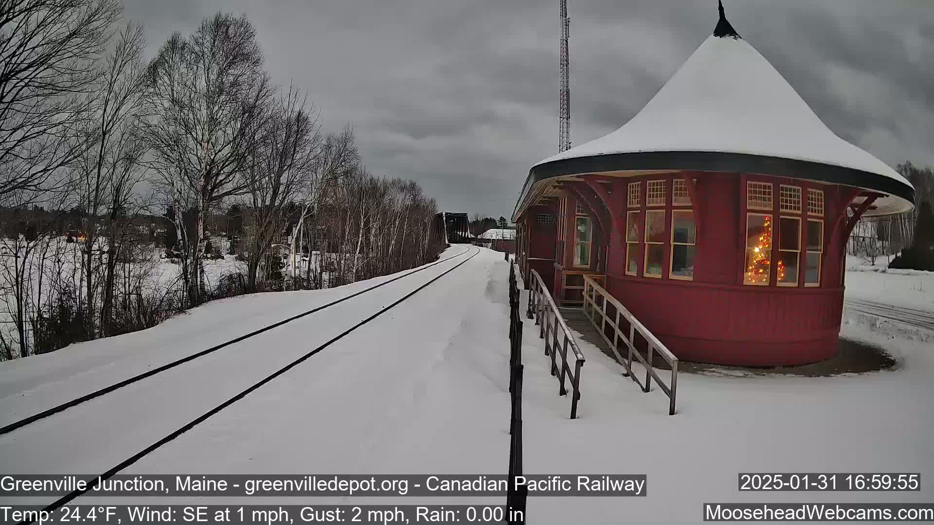 A snow-covered railroad track leads to a red, round train station with a lit Christmas tree visible in the window, under an overcast sky.