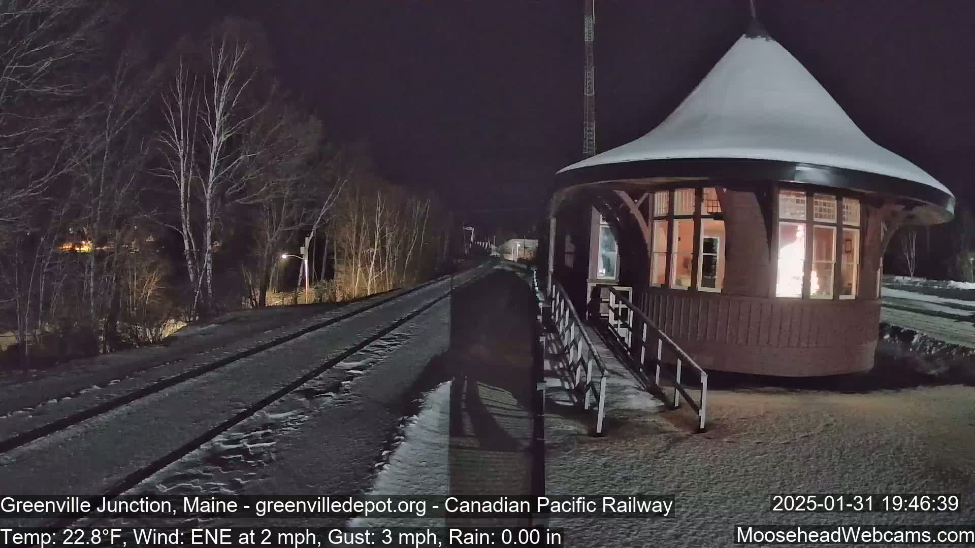 A snow-covered train station at night with a lighted interior, next to a snow-covered railway track, under a dark sky.