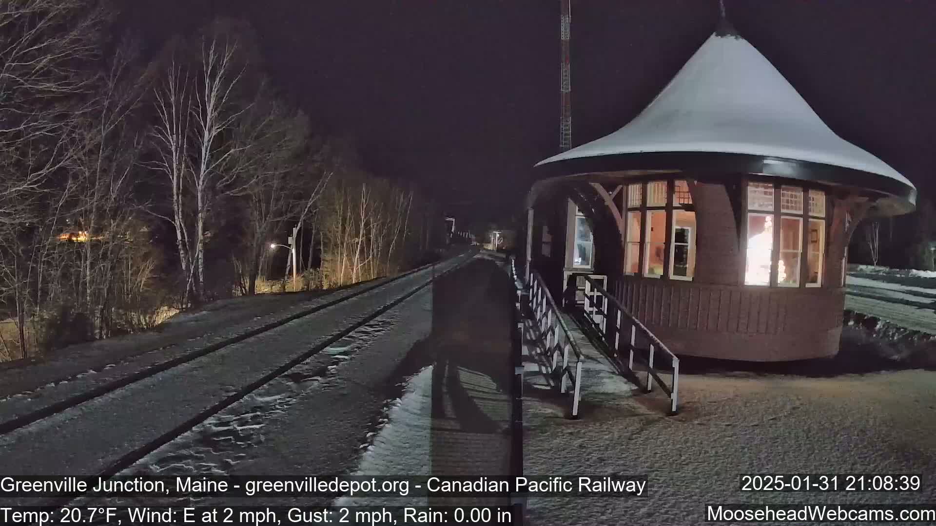 A snow-covered train platform and tracks are seen at night, with a small, snow-covered building illuminated from within.