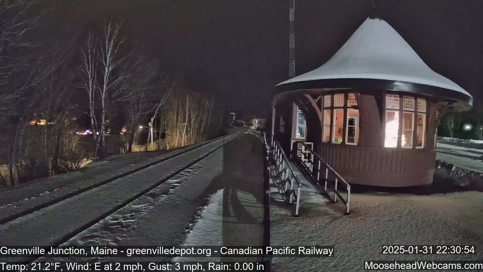 A snow-covered, round train depot with lit windows is situated beside a snow-dusted railway track at night, under a dark sky with an easterly wind.