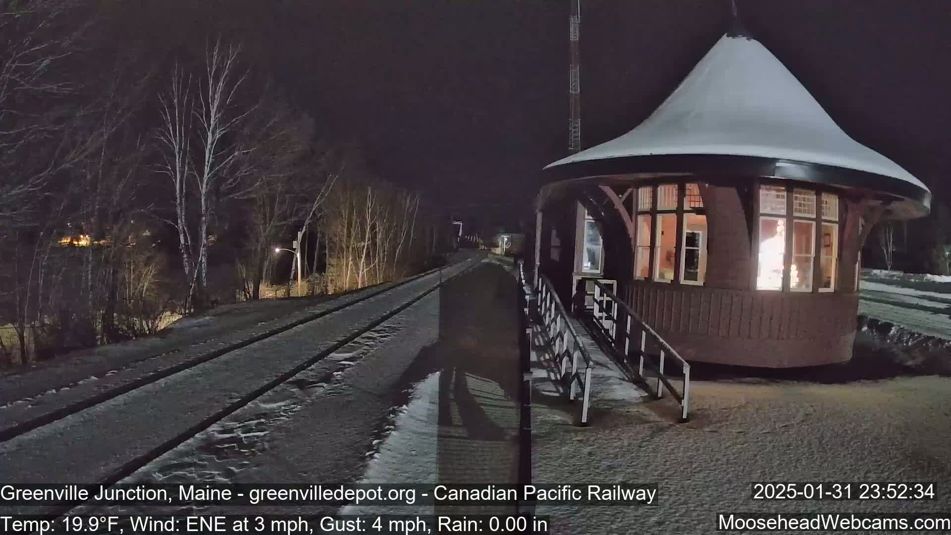 A snow-covered, illuminated train station stands beside a set of train tracks at night.