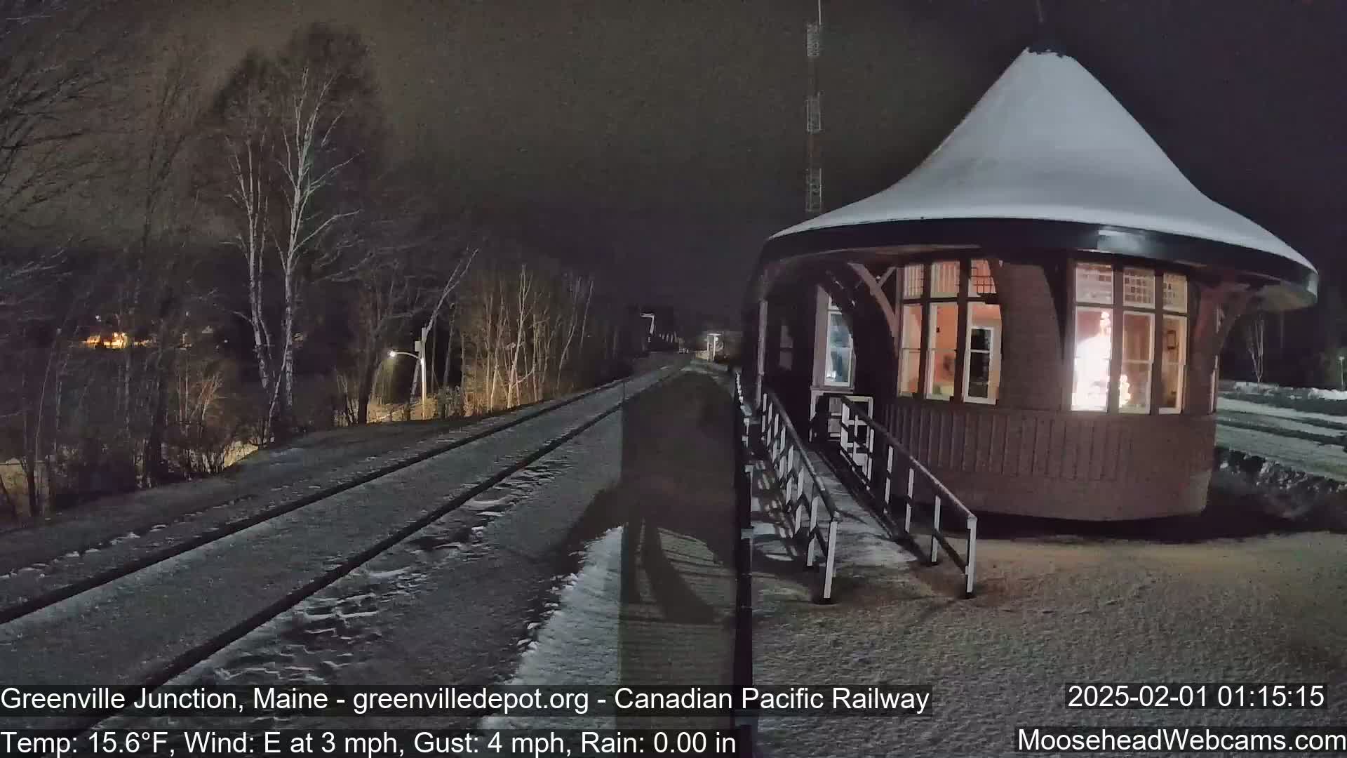 A snow-covered train depot with a lit interior is situated beside snow-covered train tracks at night under a dark sky.