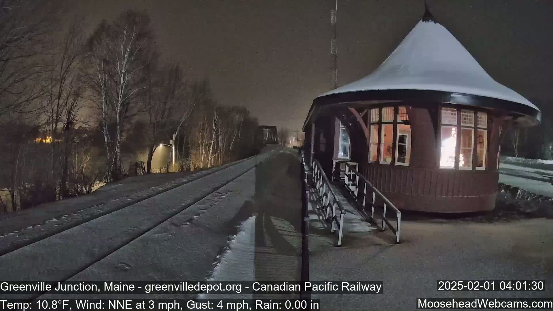 A snow-covered train platform and tracks are illuminated at night, next to a small, round, snow-dusted building with a lit window.