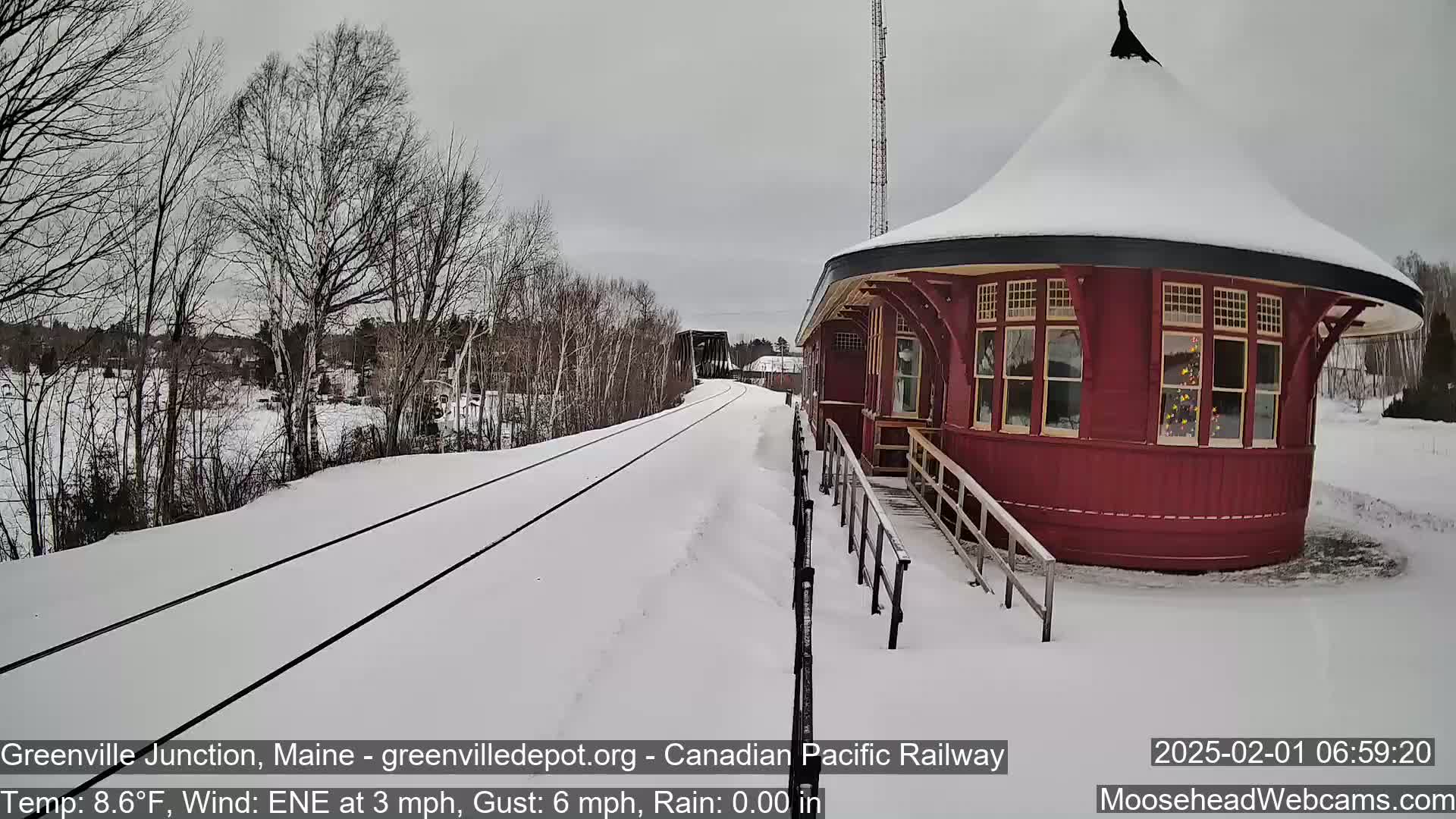 A snow-covered railroad track leads to a round, red train depot under a gray sky.