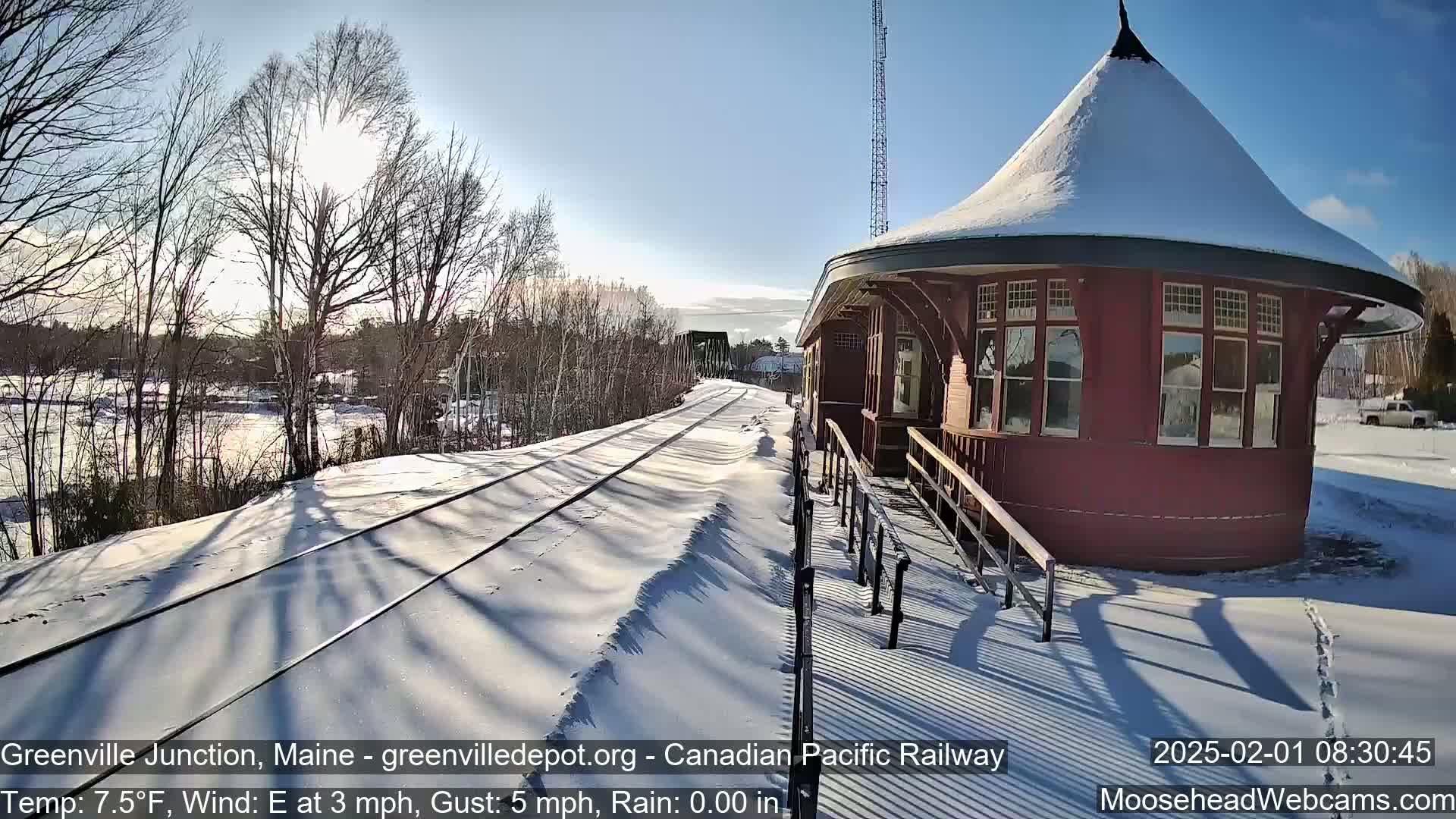 A snow-covered train platform with a round, red train station sits beside a set of train tracks on a sunny day with a light east wind.