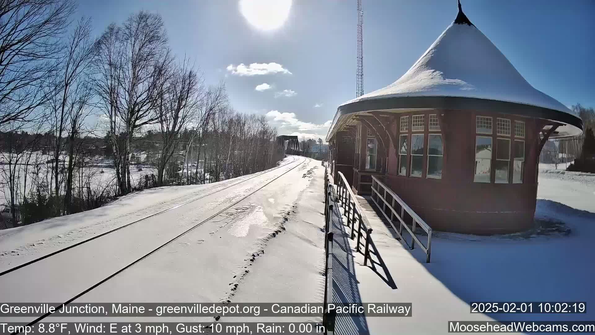 A snow-covered train platform with a red, snow-topped building stands beside a set of train tracks on a sunny day with a light east wind.
