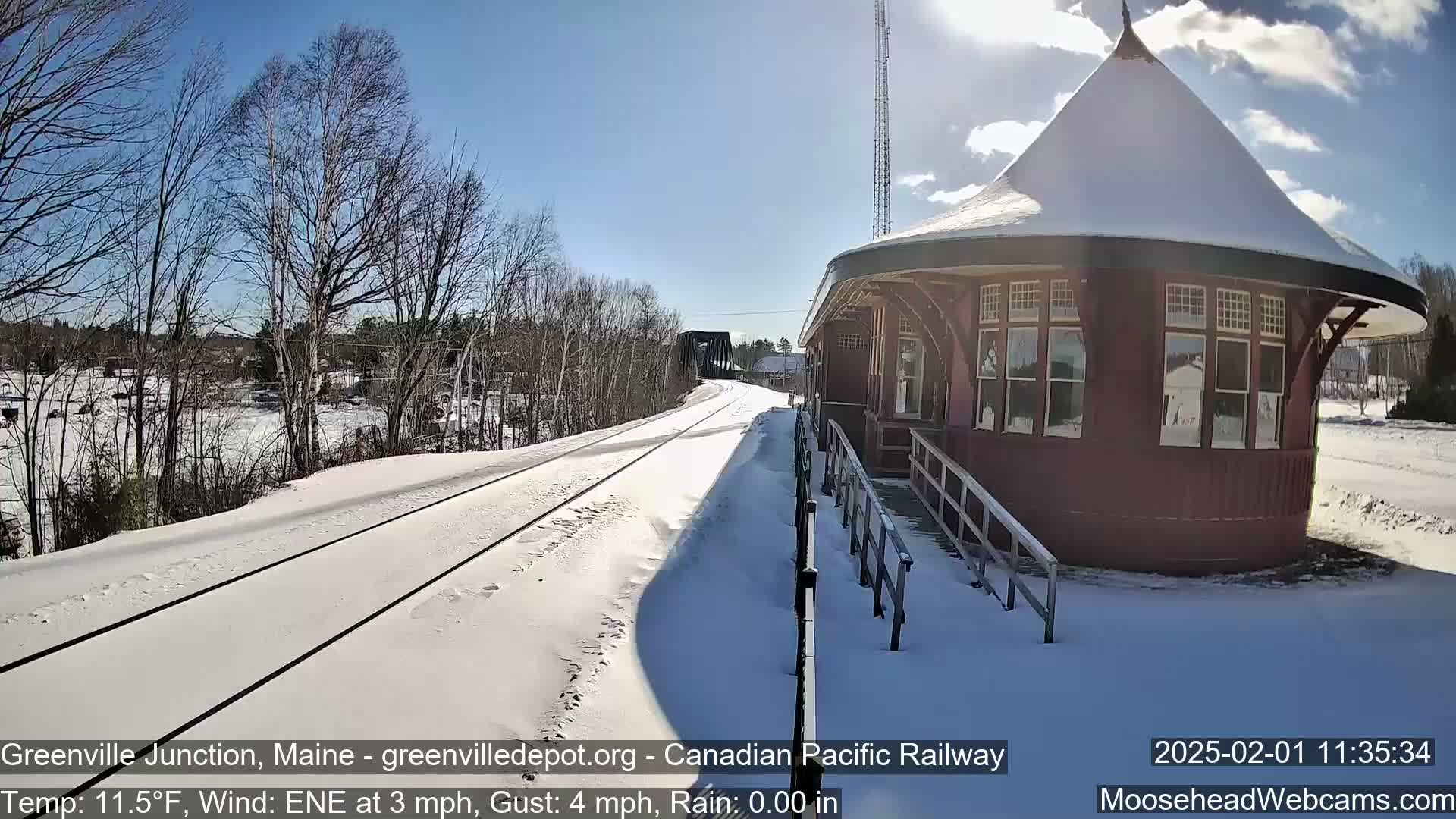 A snow-covered train track leads to a snow-covered, octagonal train station under a sunny sky.