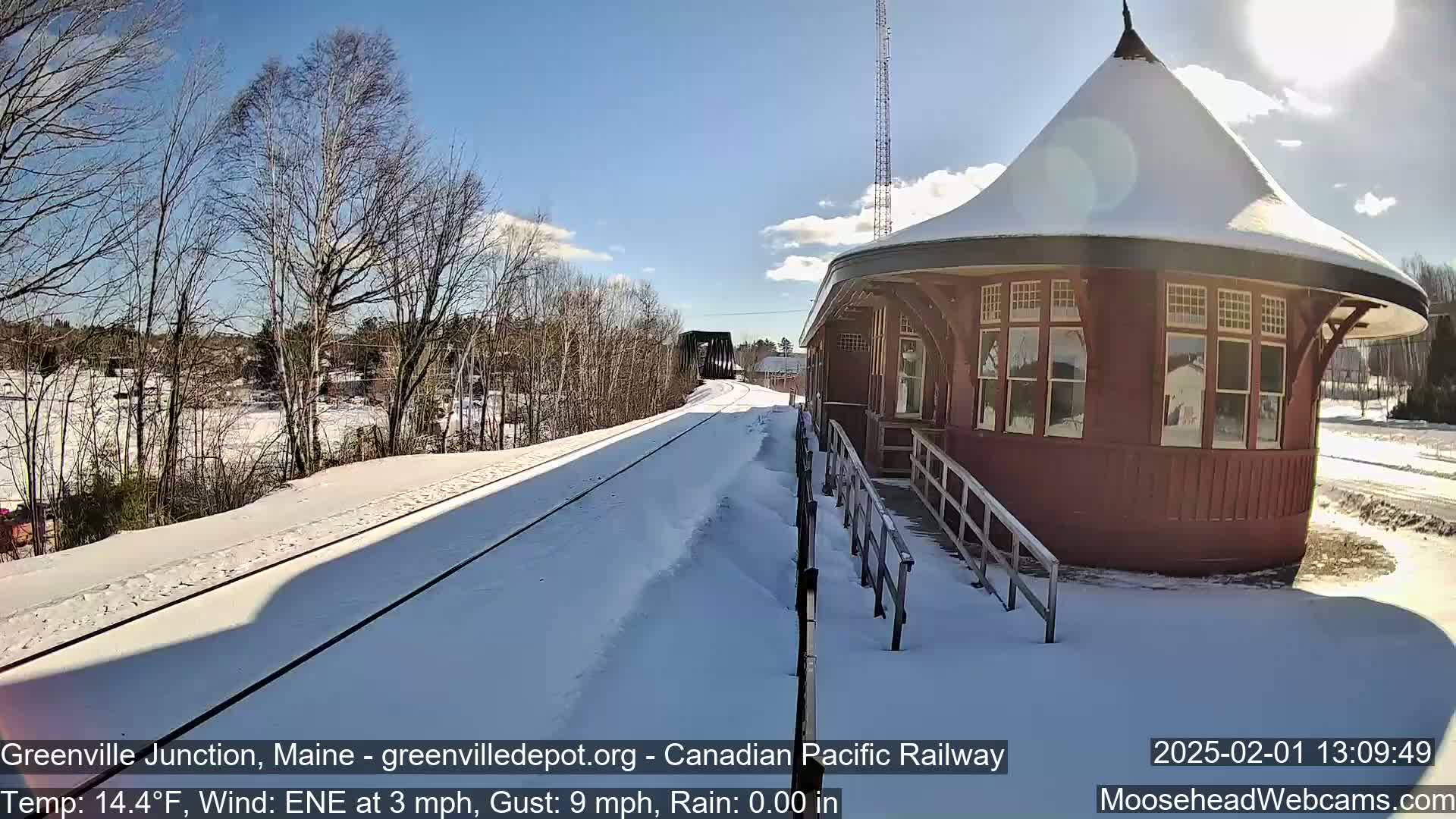 A snow-covered railway track leads to a red, round train station under a sunny sky.