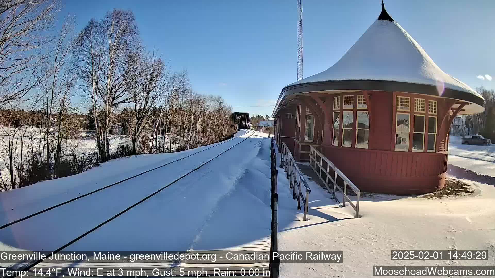A snow-covered train station with a round, red roof sits next to a set of train tracks on a sunny day with a light east wind.