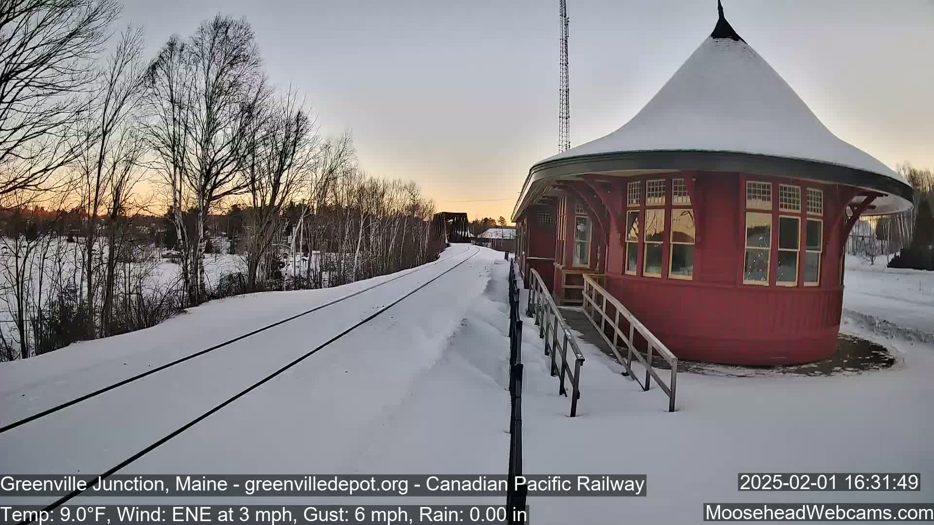 A snow-covered railway track extends towards a red, round train station at sunset.