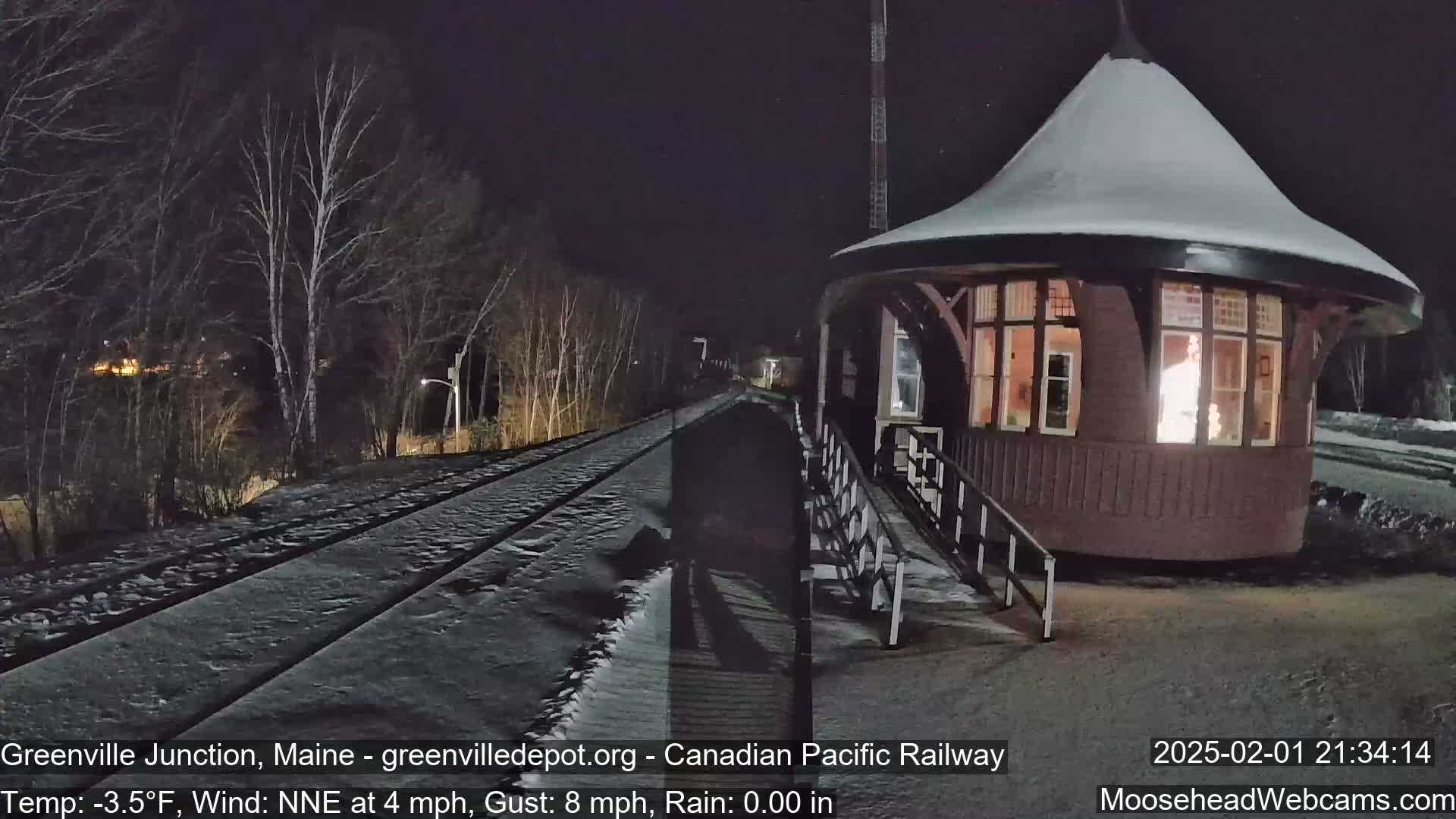 A snow-covered railway track at night passes a lit-up, snow-covered, round train station under a dark, clear sky with a slight wind.