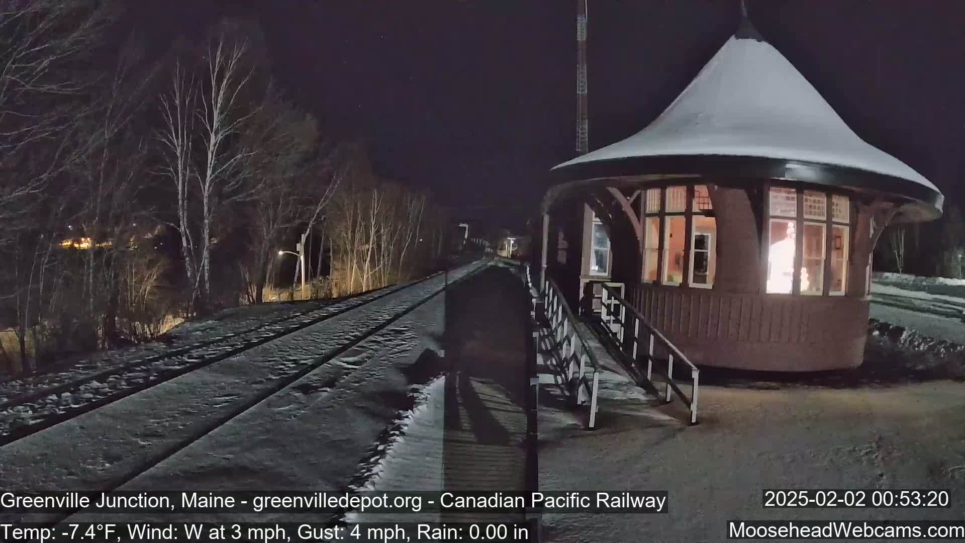 A snow-covered train station with a lit interior stands beside a snow-covered railway track at night, under a dark sky with a few stars visible.