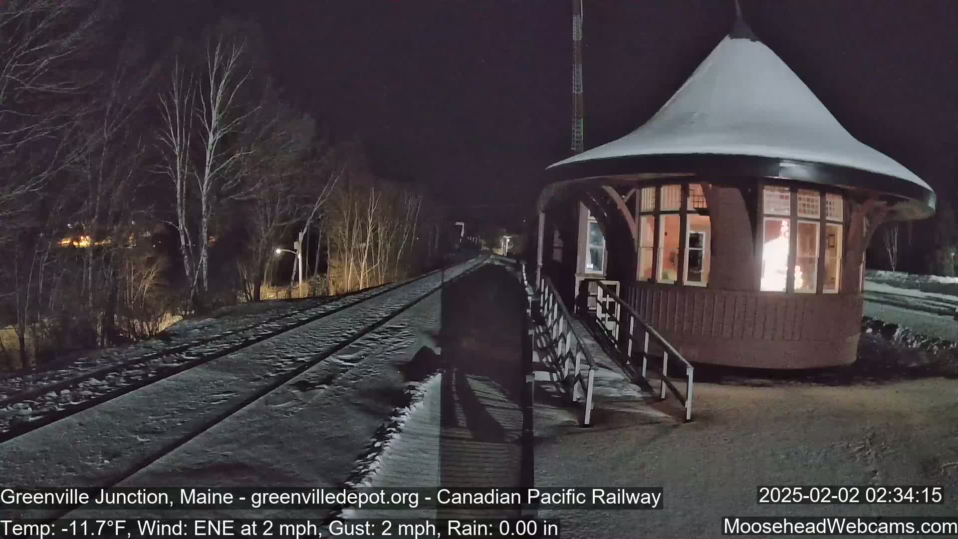 A snow-covered train platform and tracks are illuminated at night next to a small, lit-up building.