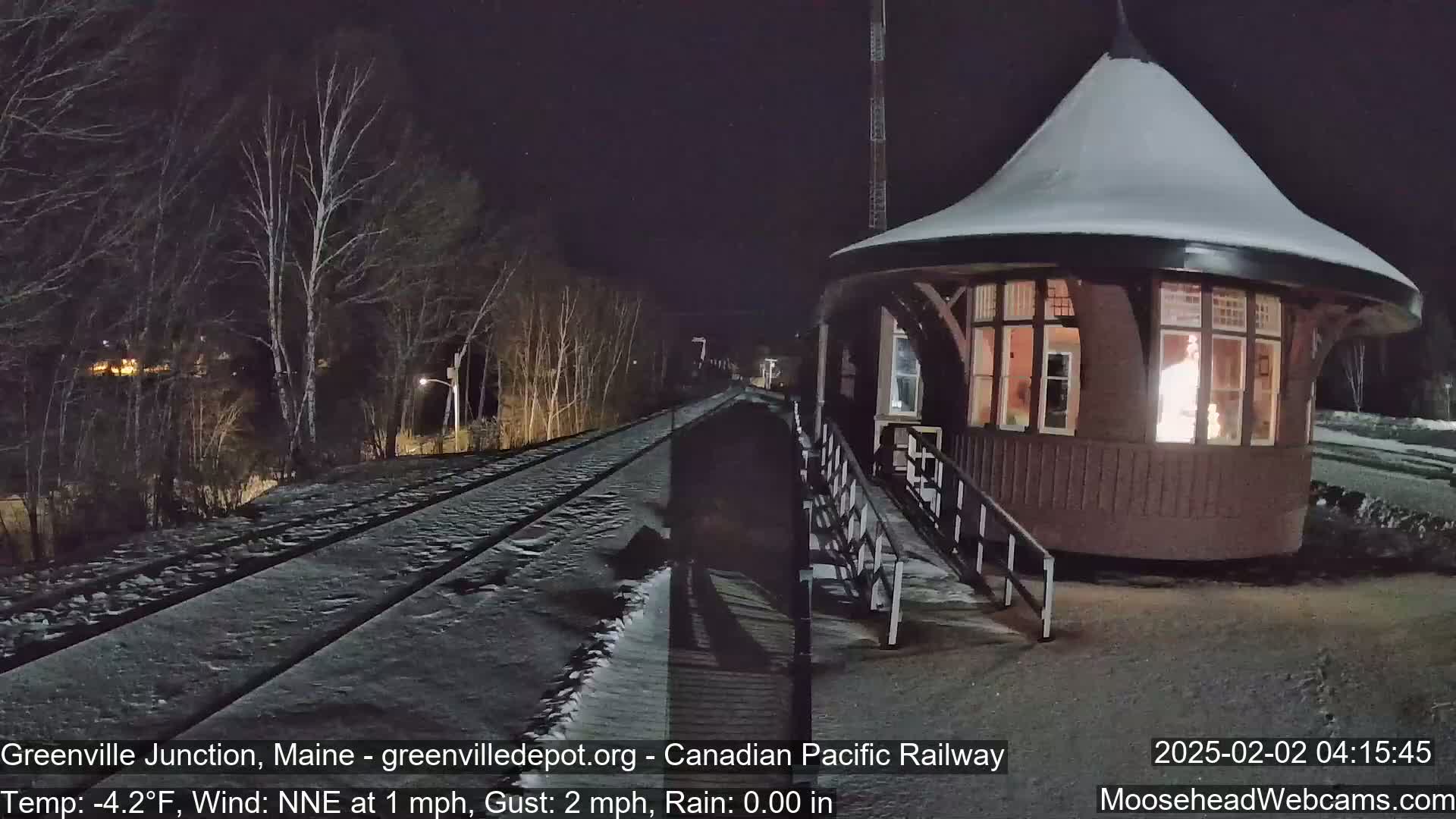 A snow-covered train platform and tracks are illuminated at night, next to a small, snow-covered, round building with a lit interior.
