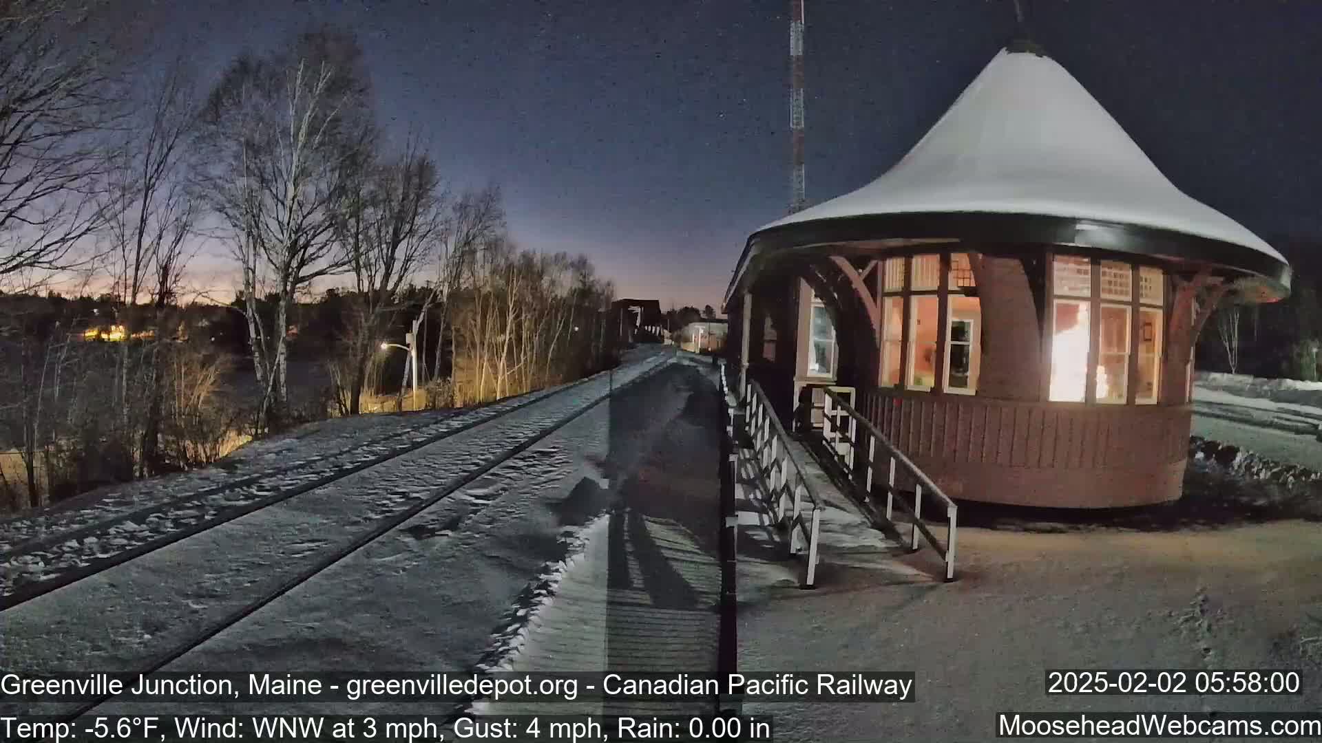 A snow-covered train platform and tracks are visible under a dark night sky next to a round, snow-capped building with lit windows.