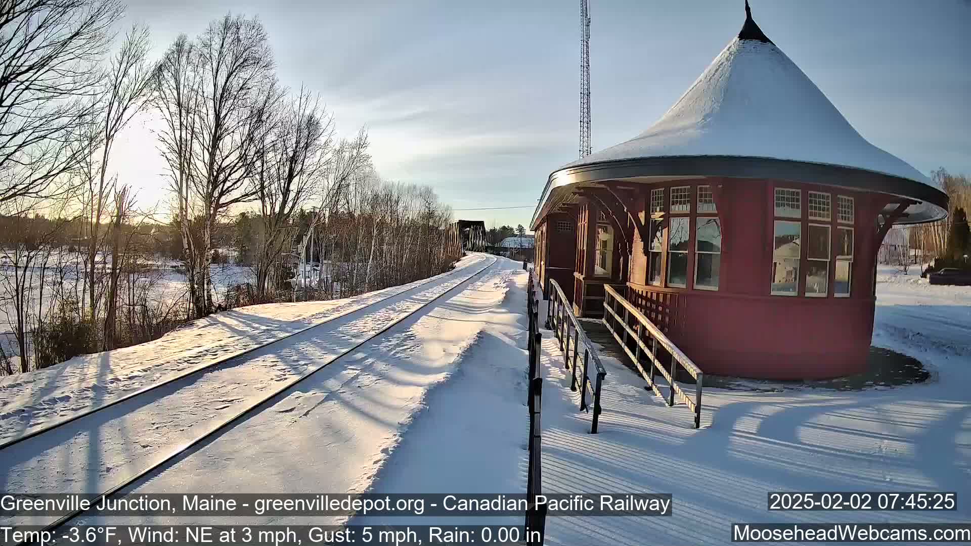 A snow-covered railway track leads to a red, octagonal train station under a mostly clear, sunny sky.