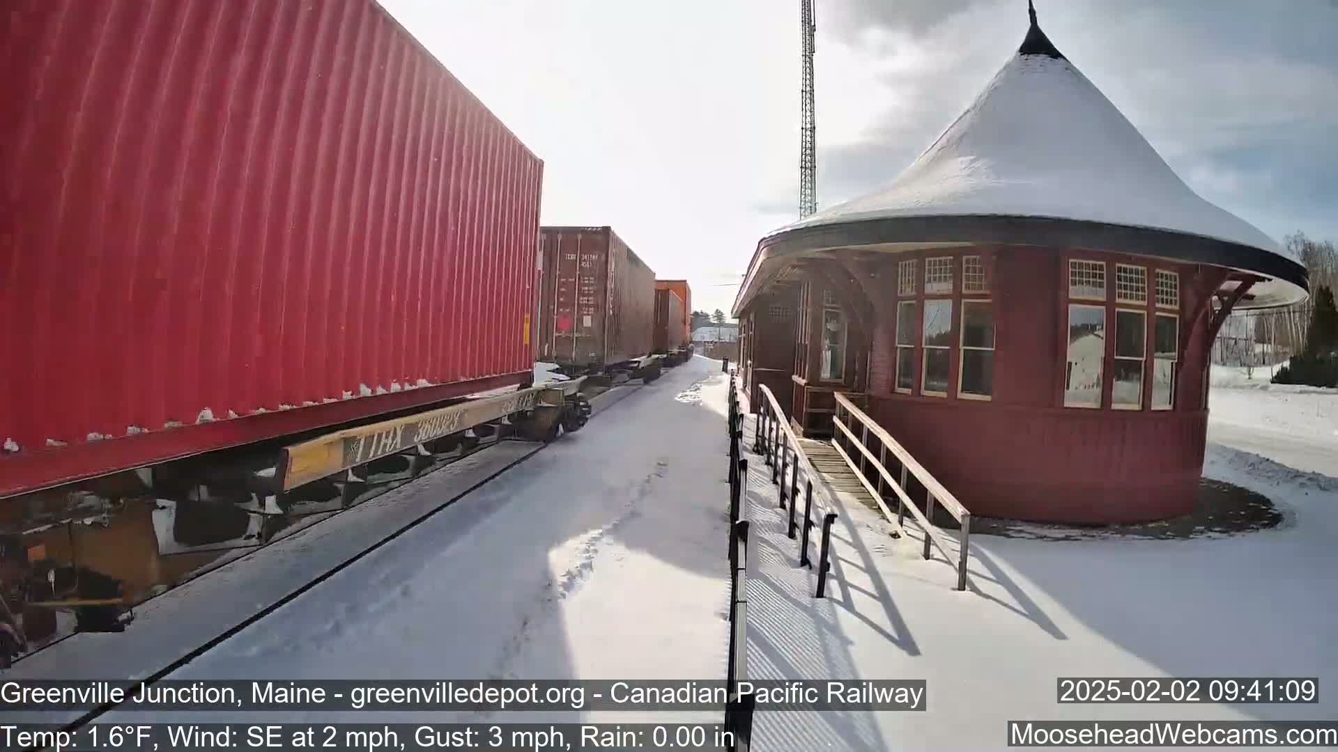 A red freight train sits partially in the snow next to a snow-covered, round red train station on a sunny day.
