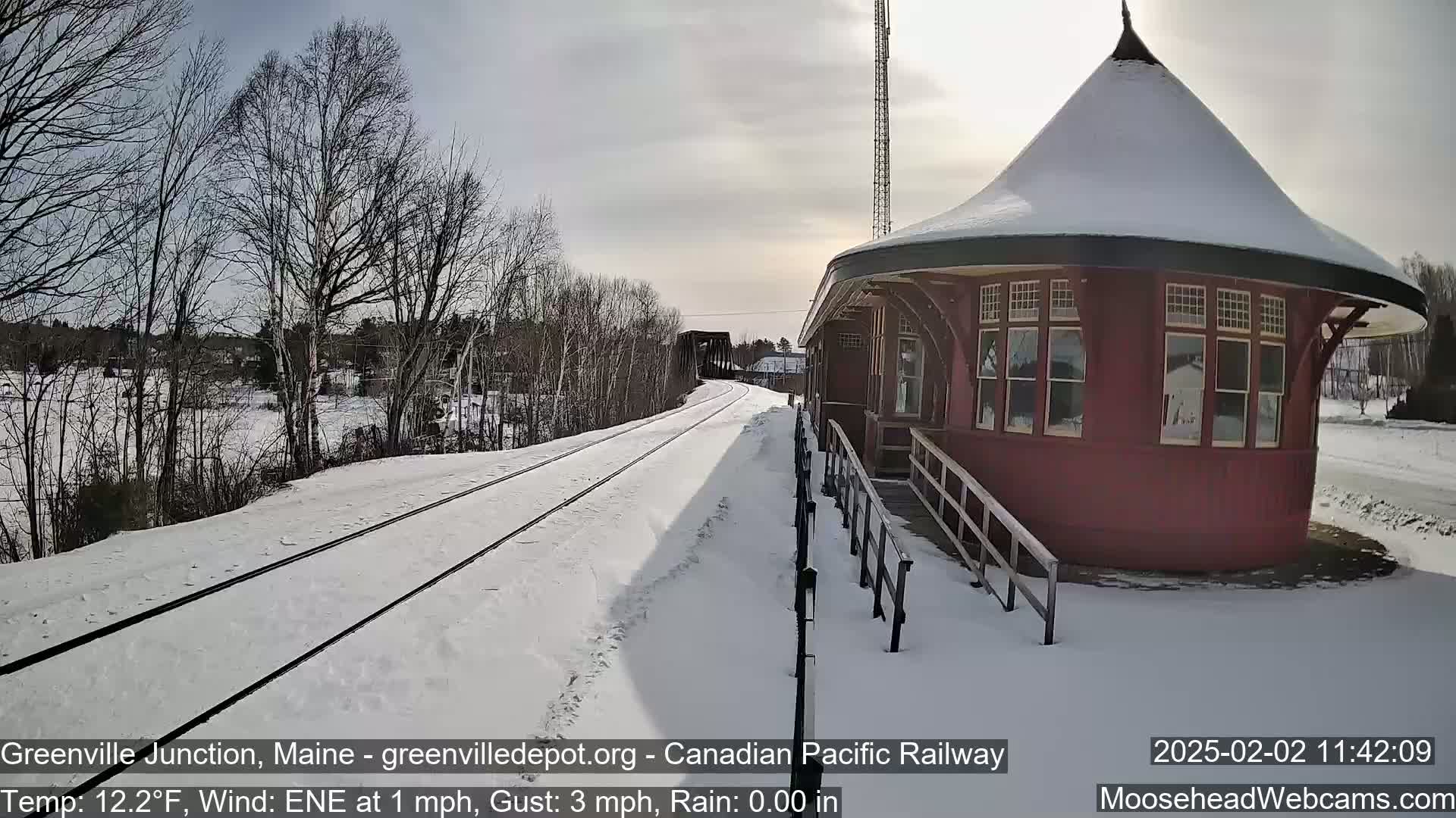 A snow-covered railway track leads to a red, octagonal train station under a partly cloudy sky.