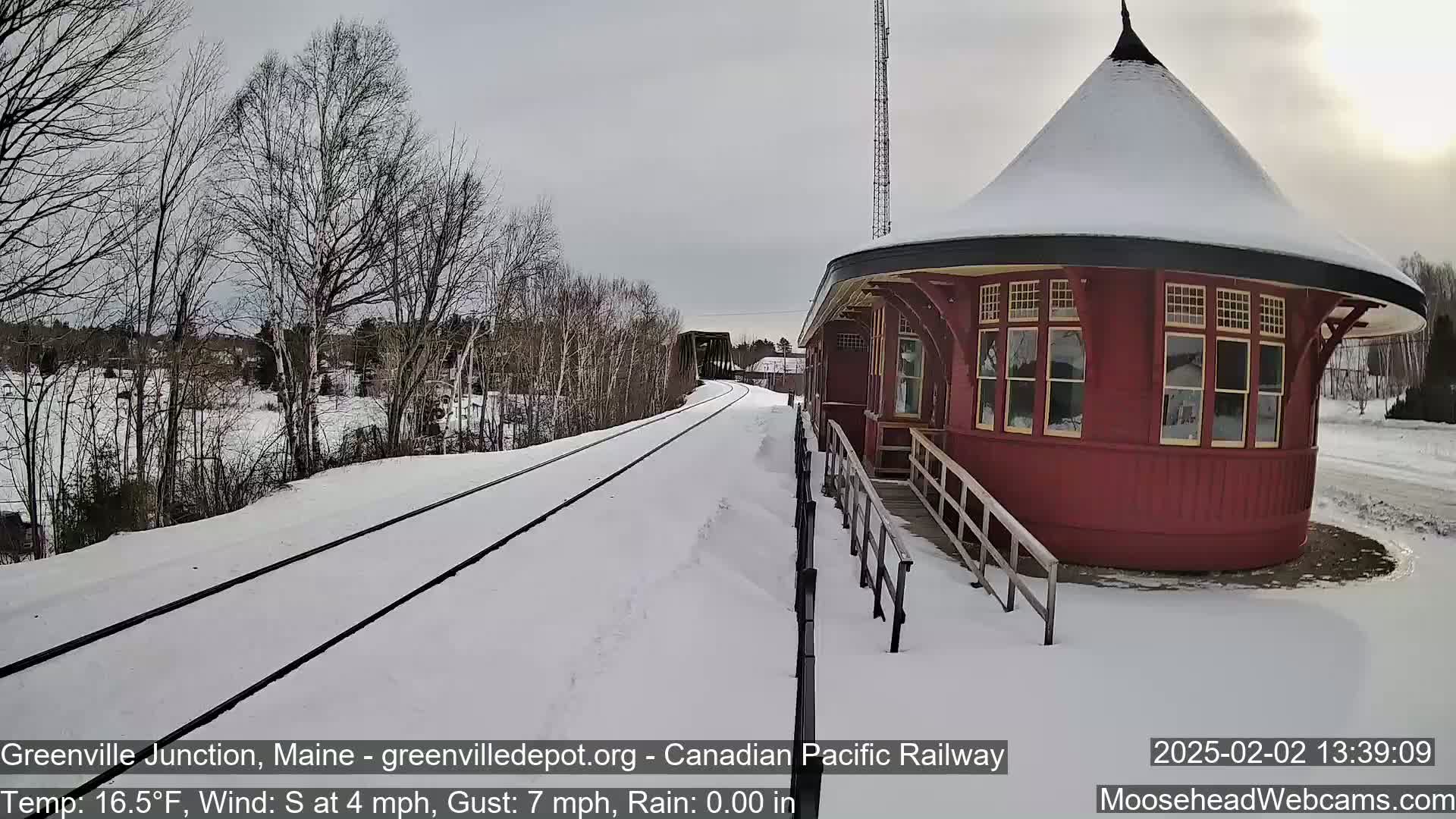 A snow-covered railway track leads to a red, round train depot under a cloudy sky.