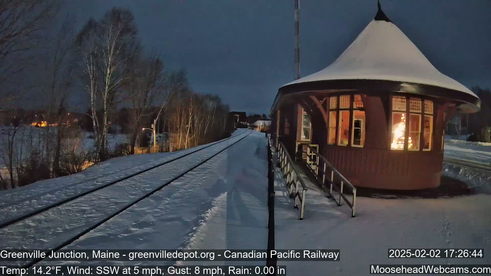 A snow-covered train station platform with a round, snow-topped building and railroad tracks stretches into the twilight.