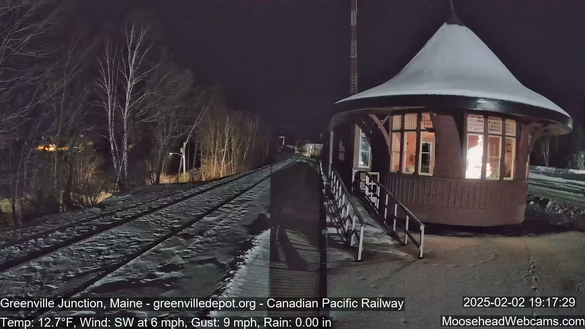 A snow-covered railway track leads to a lit-up, octagonal, wooden train station at night under a dark sky.