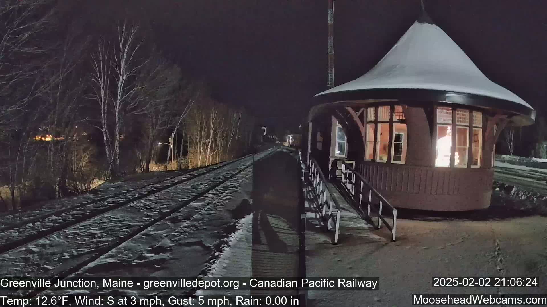 A snow-covered train track leads to a lit-up, snow-dusted train station at night under a dark sky.