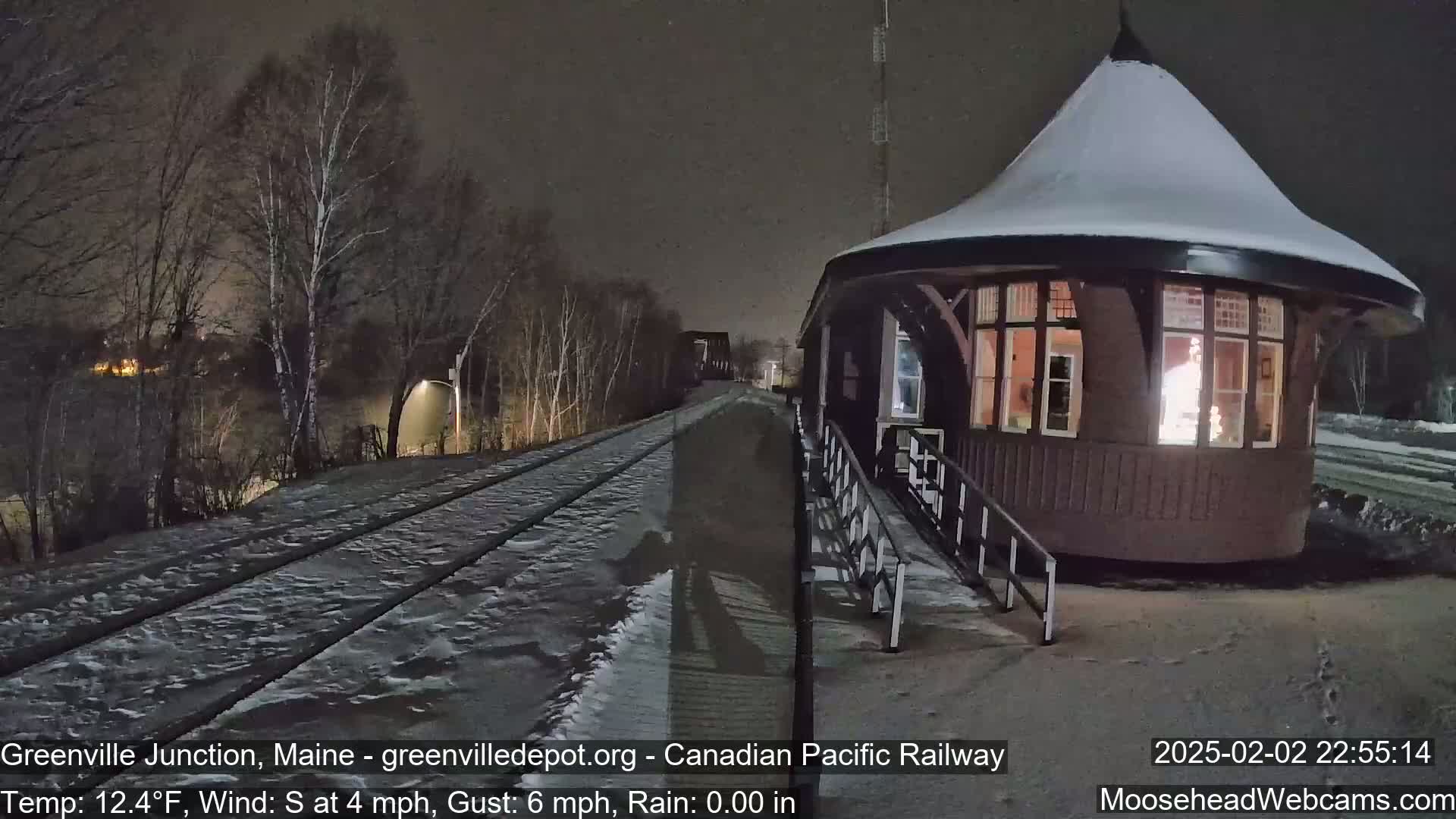 A snow-covered train platform with a small, illuminated, snow-capped building next to a set of train tracks at night.
