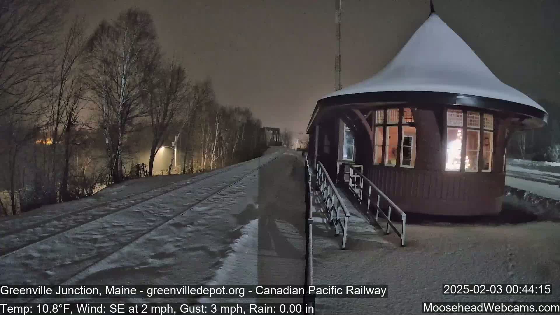 A snow-covered train station with a lit interior stands beside a snow-covered railway track at night under a dark sky.
