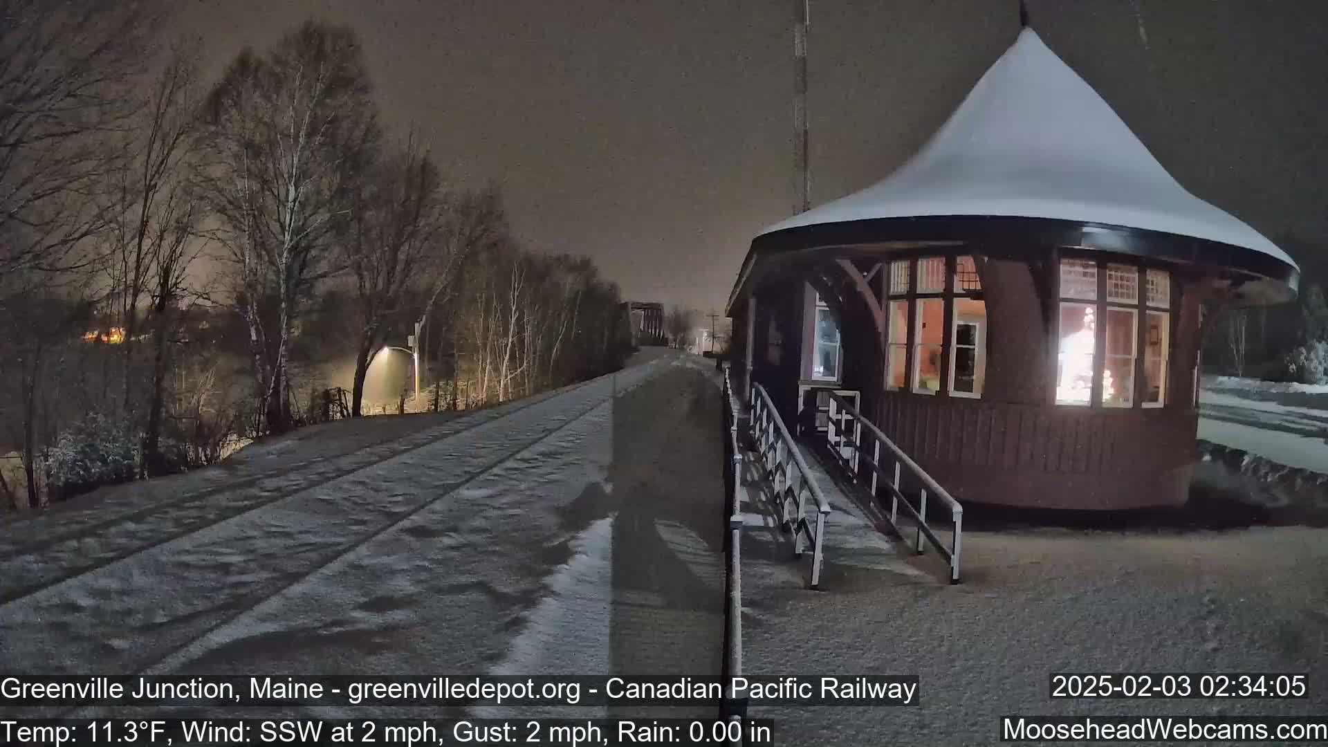 A snow-covered train depot with a lit window is visible at night, with lightly falling snow and a rail line extending into the background under a dark sky.
