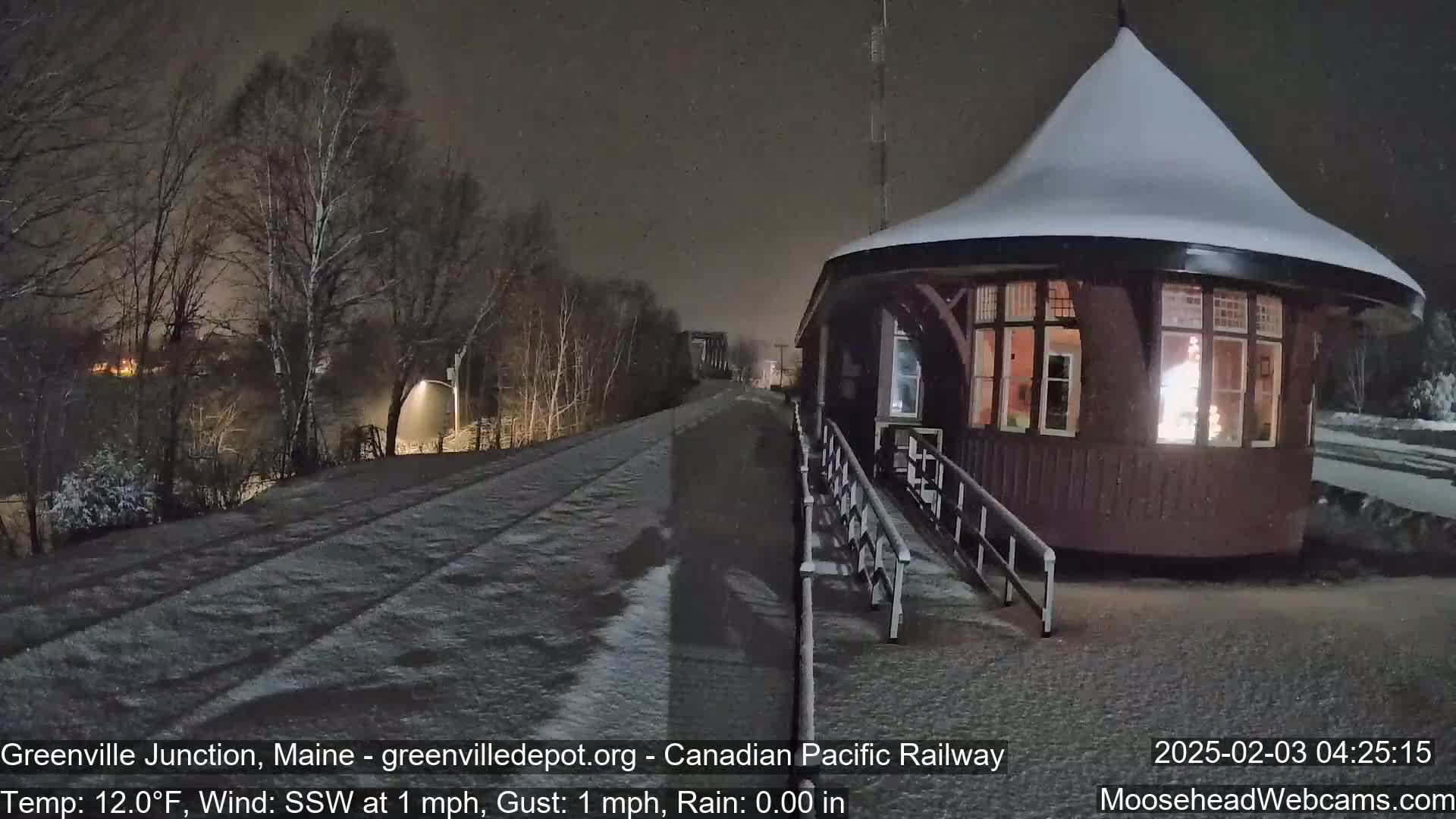 A snow-covered train depot with a lit interior is visible at night, under light snowfall with a calm south-southwesterly wind.