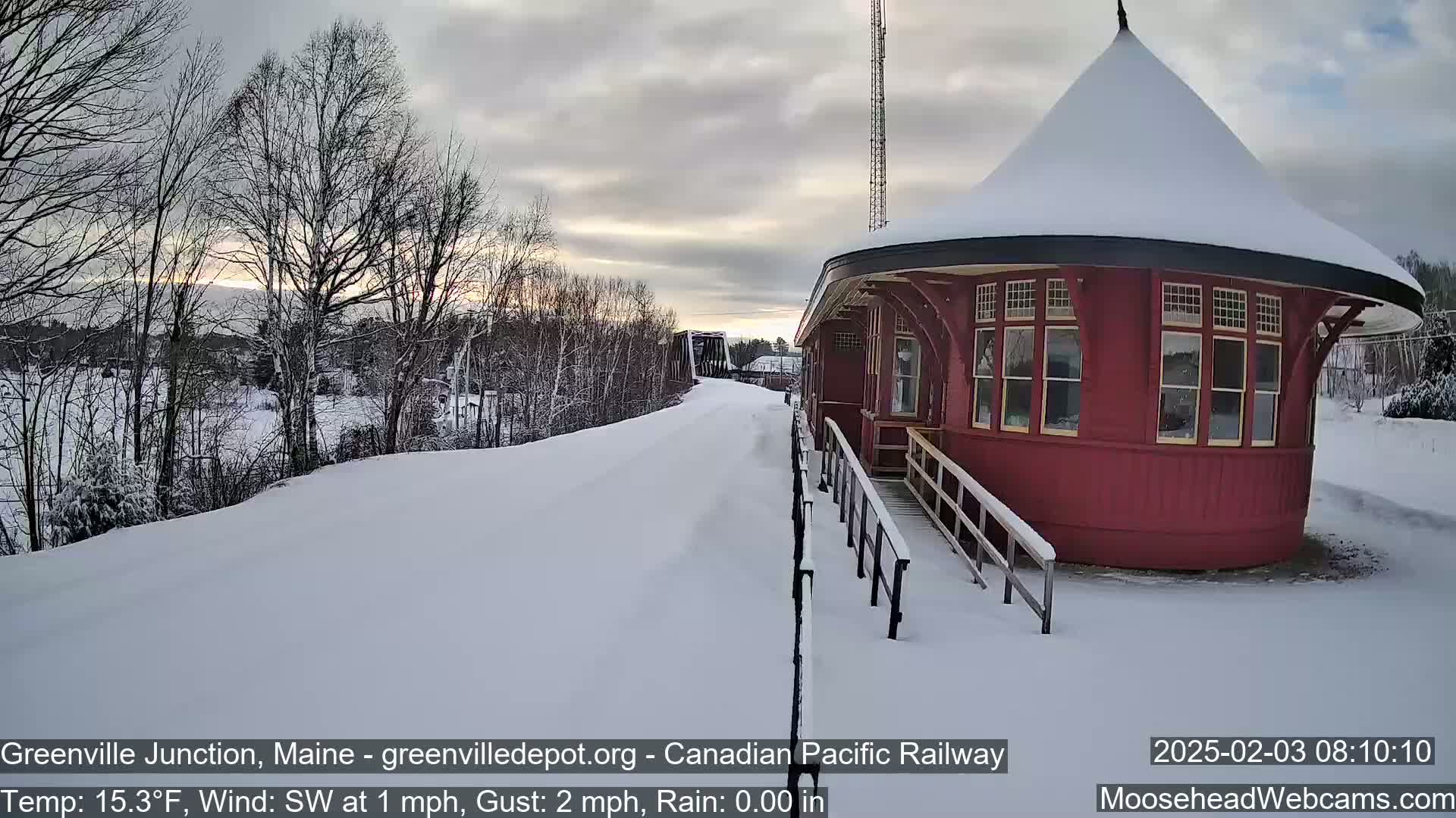 A snow-covered train platform with a red, round train station is visible under a partly cloudy sky.