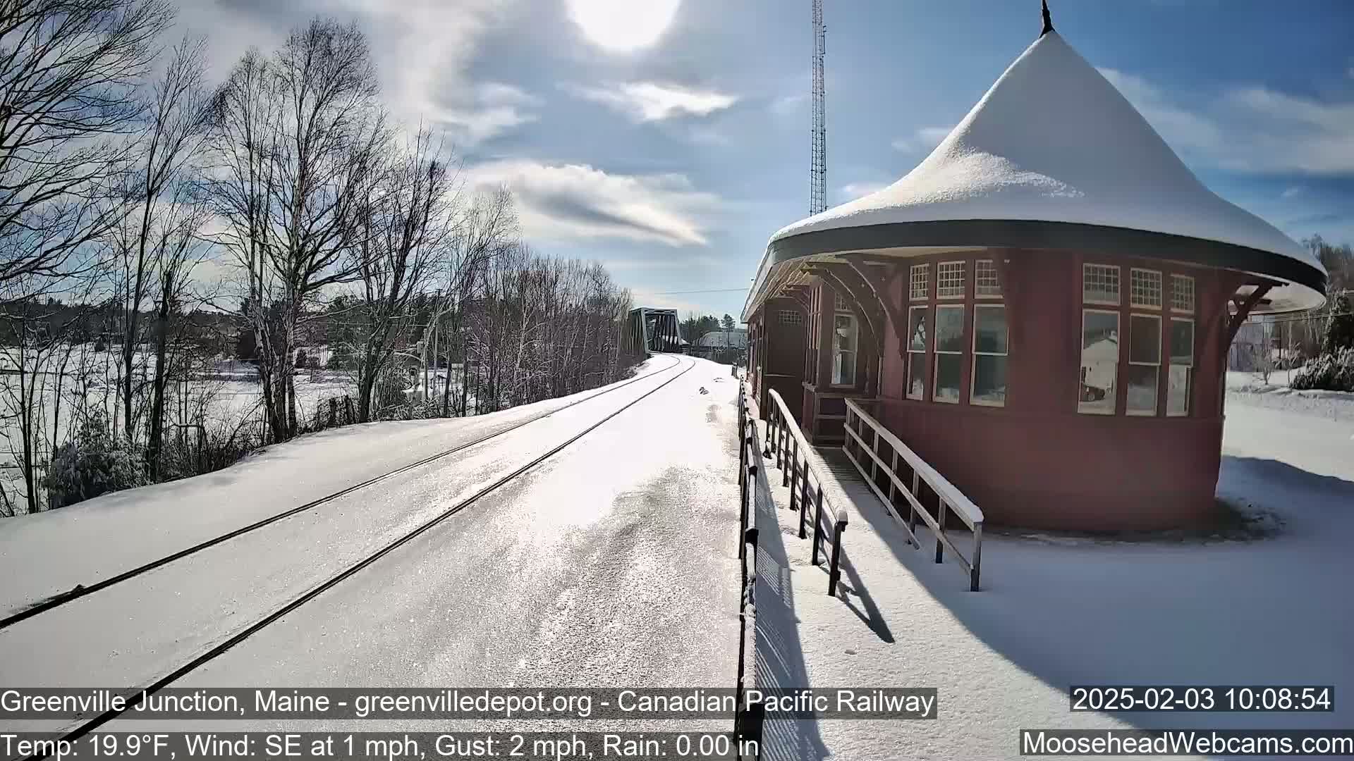 A snow-covered train platform and tracks extend towards a bridge under a mostly sunny sky.