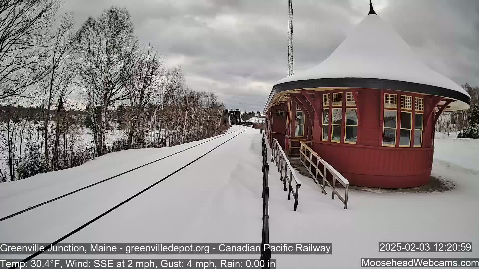 Snow-covered train tracks lead to a red, round train station under a gray, cloudy sky.