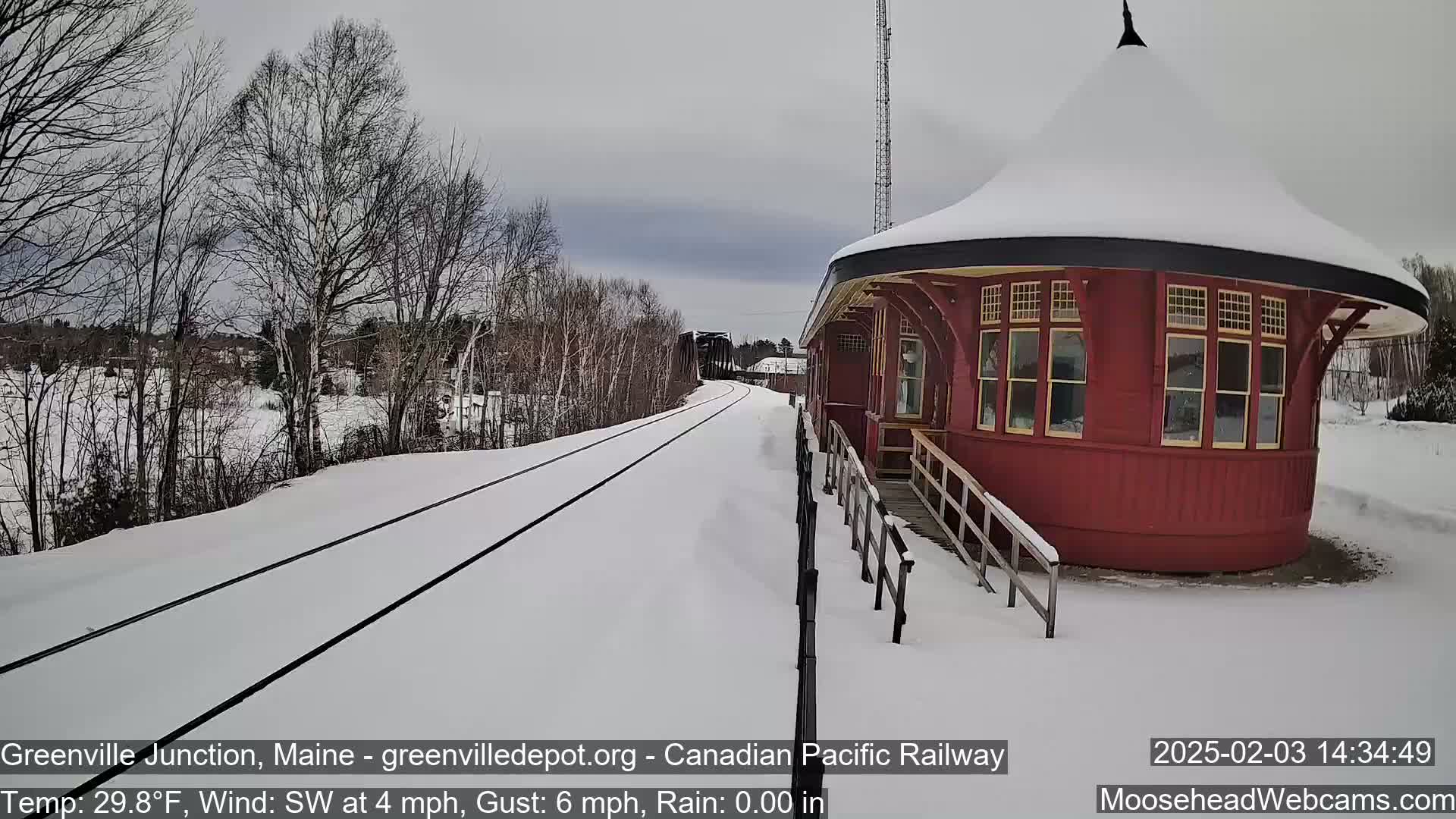 Snow-covered railroad tracks extend toward a red, round train depot under a cloudy sky, with bare trees lining the tracks.