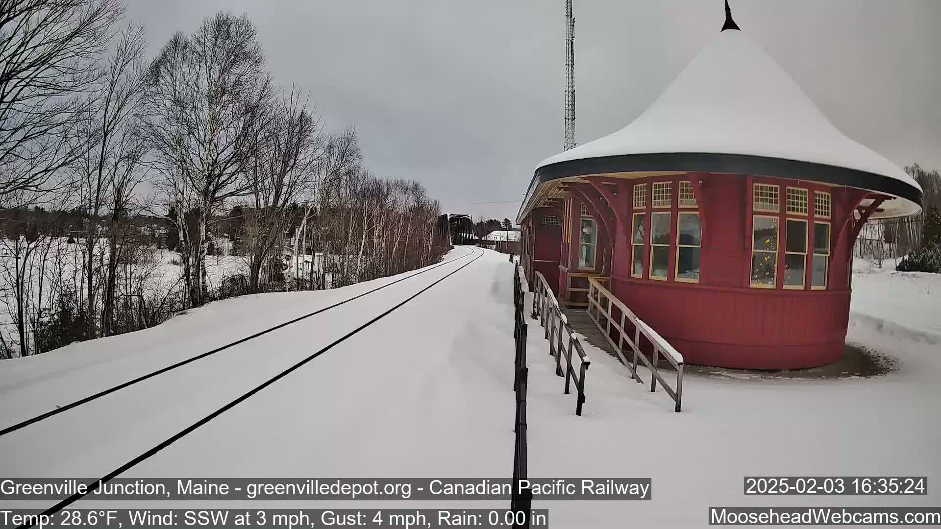 A snow-covered train platform with a red round building next to train tracks under an overcast sky.