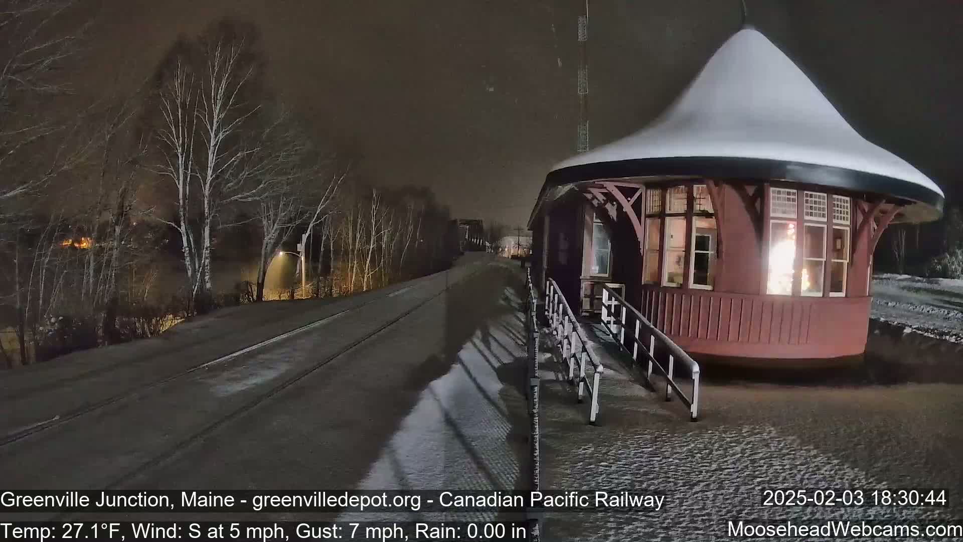 A snow-covered, red train station with illuminated windows is seen at night, next to a snow-covered road and track under a dark, snowy sky.