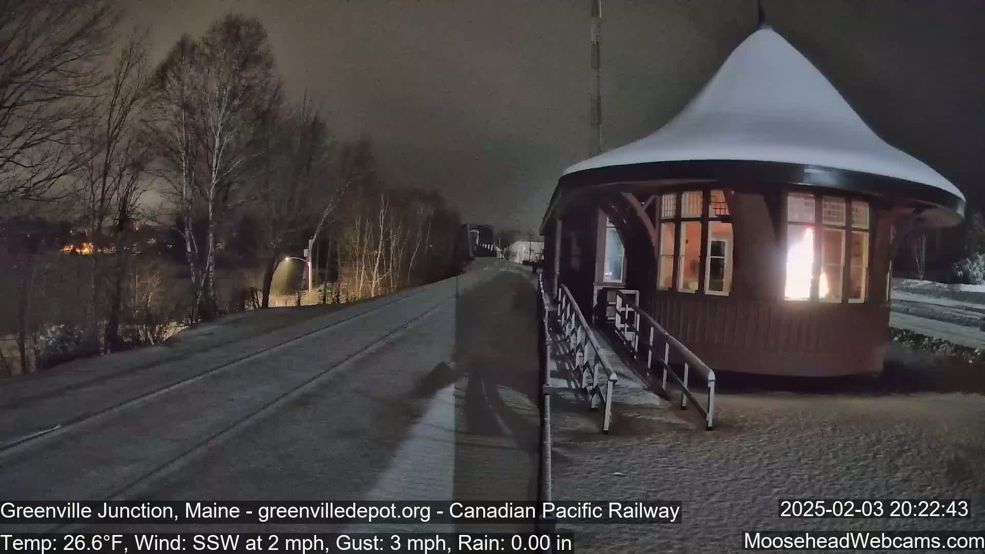 A snow-covered, dark brown railway station with a lit interior is situated beside a snow-dusted road under a nighttime sky.