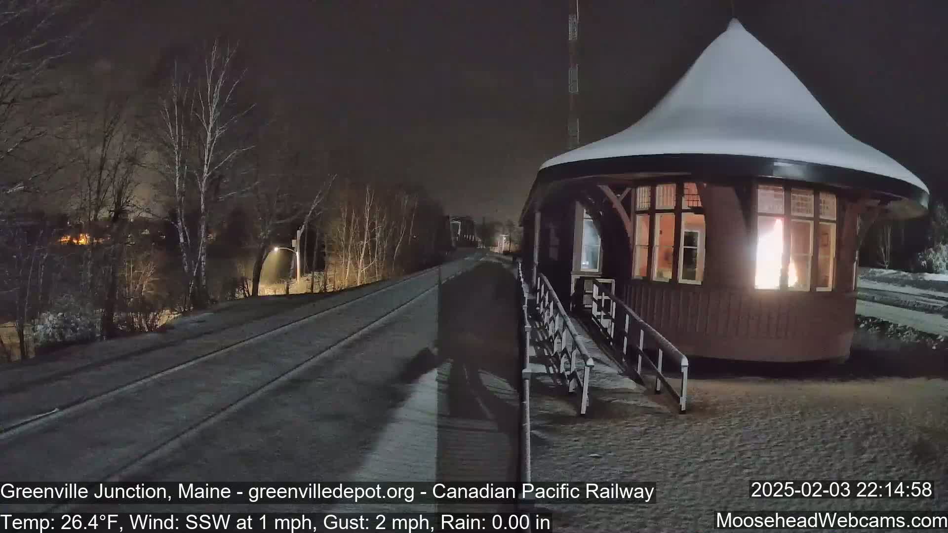 A snow-covered, illuminated train station sits beside a snow-dusted railway track at night, with bare trees and a slight wind.