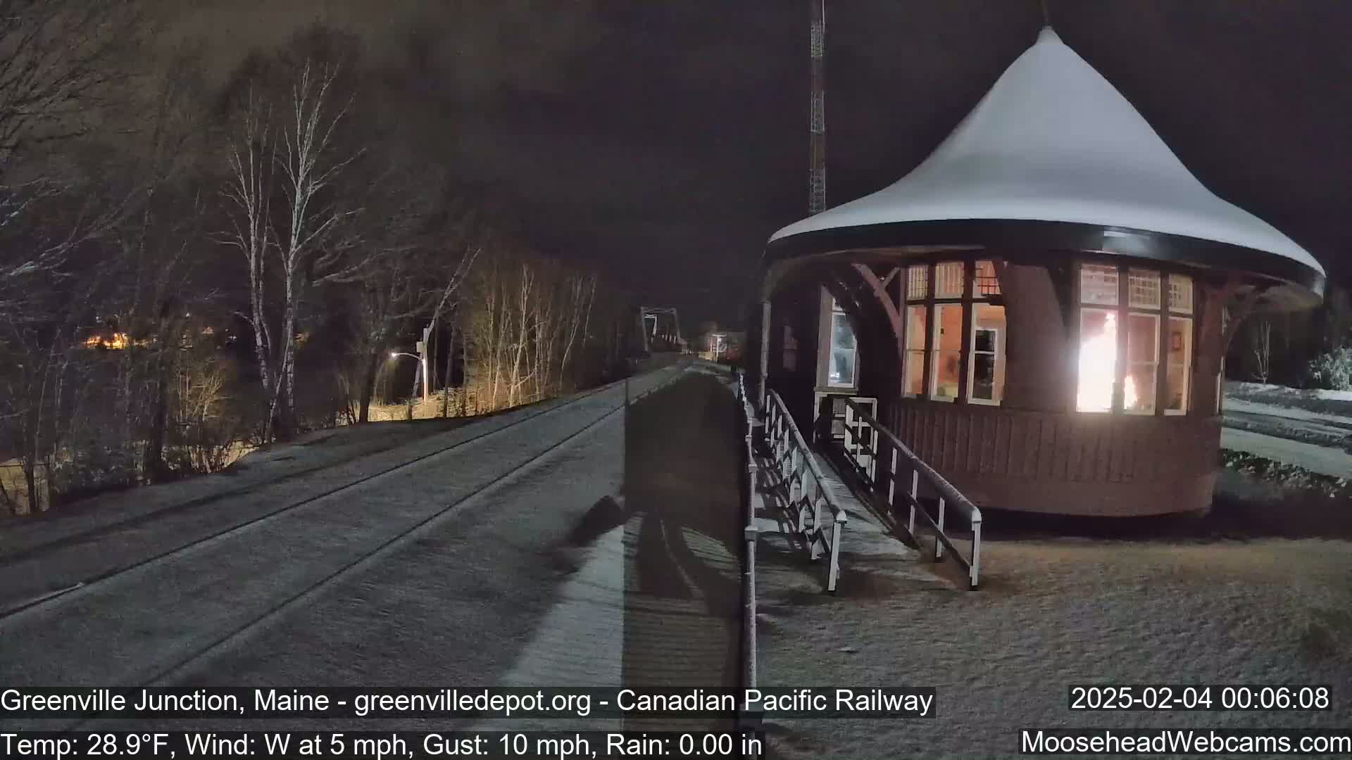 A snow-covered, dark-brown, round train station with a lit interior is situated beside a snow-dusted railway track under a nighttime sky.