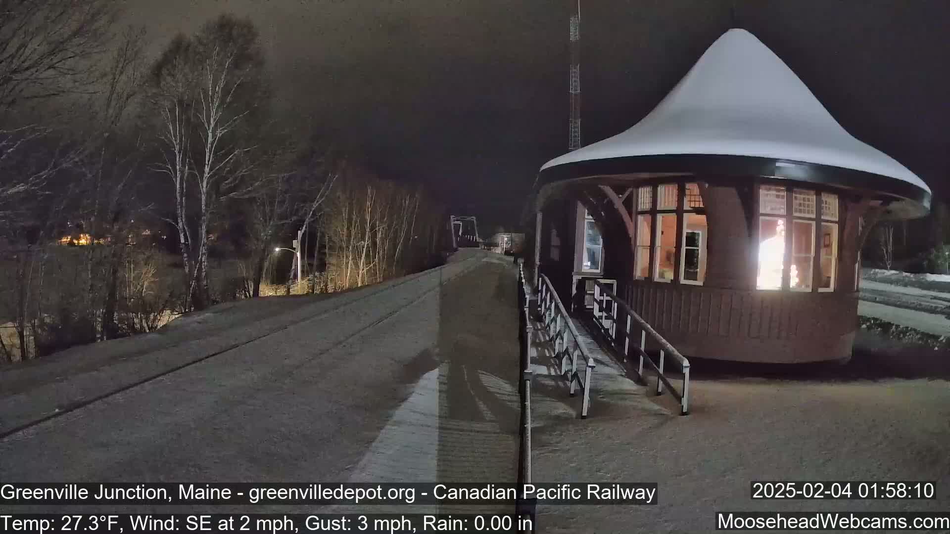 A snow-covered, illuminated train station sits beside a snow-dusted railway track at night, under a dark sky with a light southeast wind.