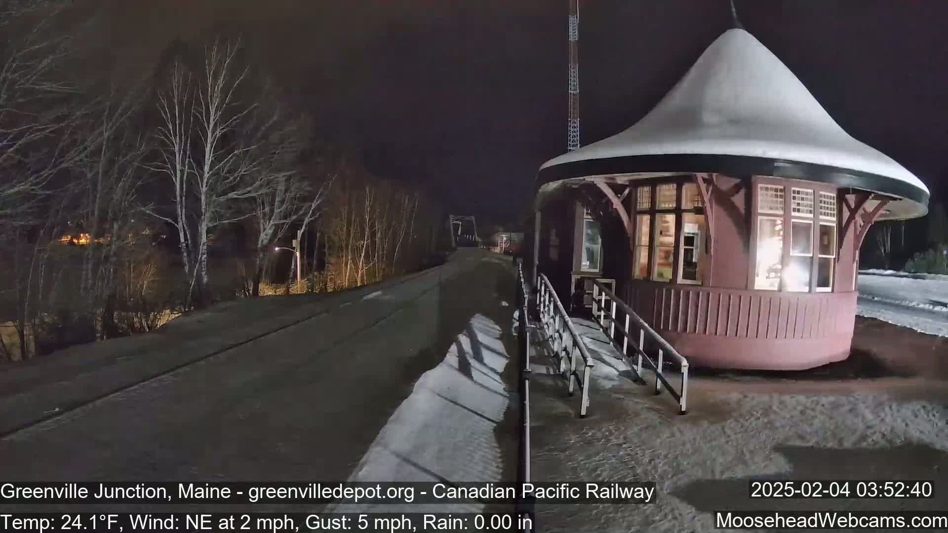 A snow-covered, round train depot stands beside a dark, snow-dusted road at night, under a clear sky with a northeast wind.