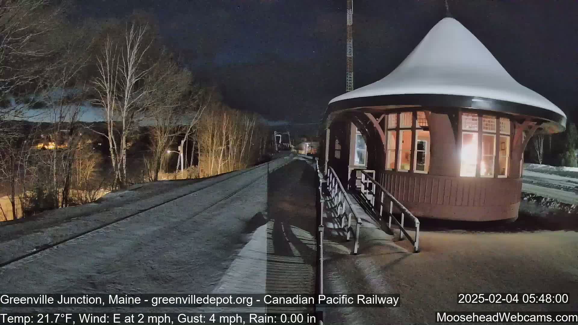 A snow-covered, illuminated train depot stands at night next to a snow-dusted railway track under a dark sky.