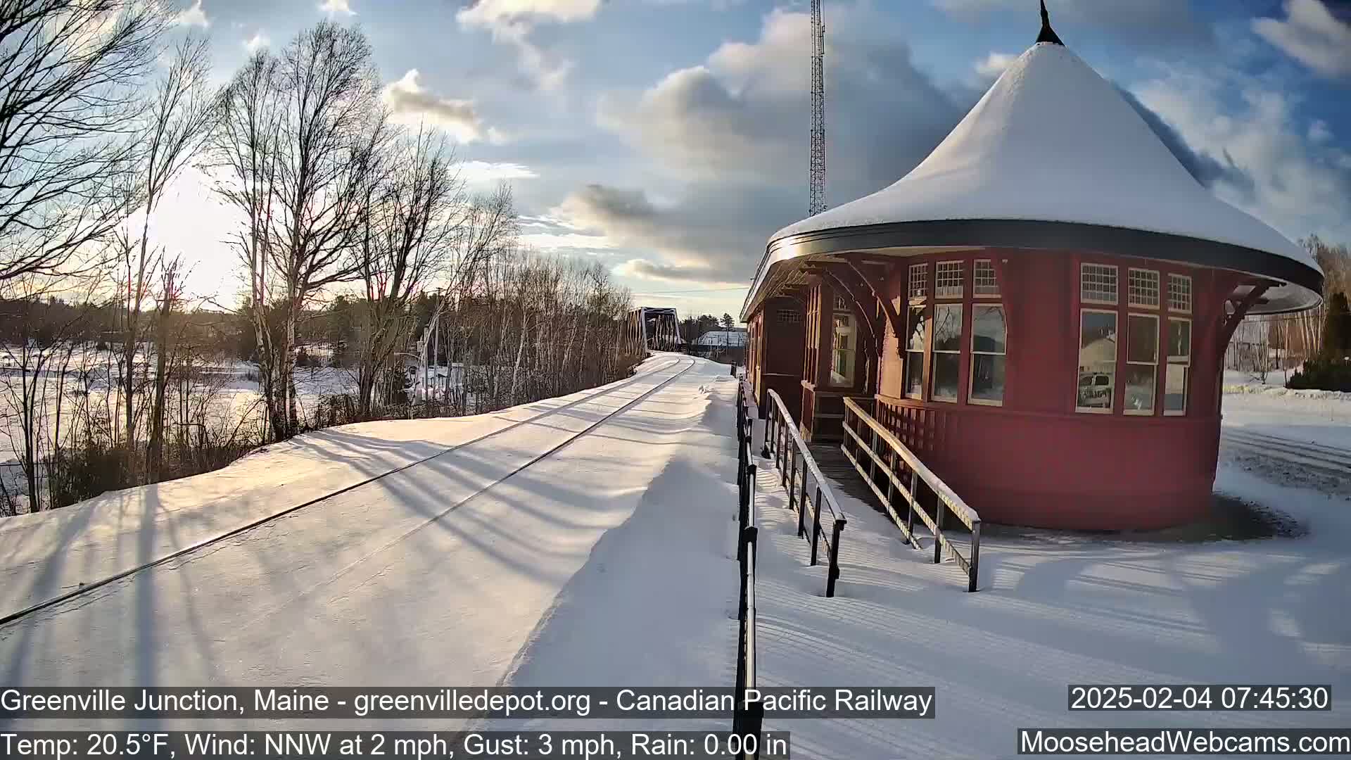 A snow-covered train platform with a red octagonal building next to a set of train tracks, under a partly sunny sky.