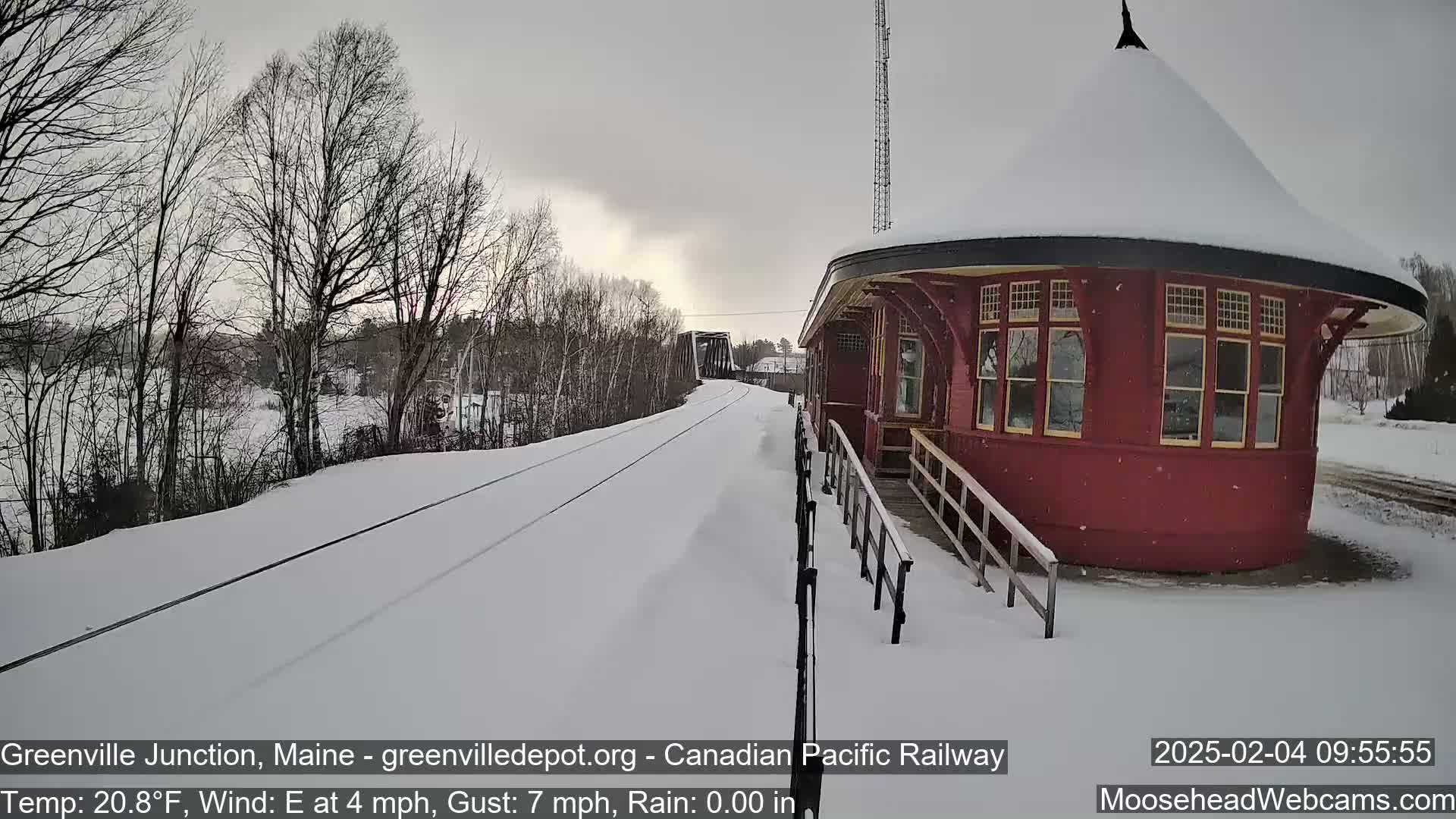 A snow-covered train platform and tracks extend toward a bridge, with bare trees and a light snowfall in the background.
