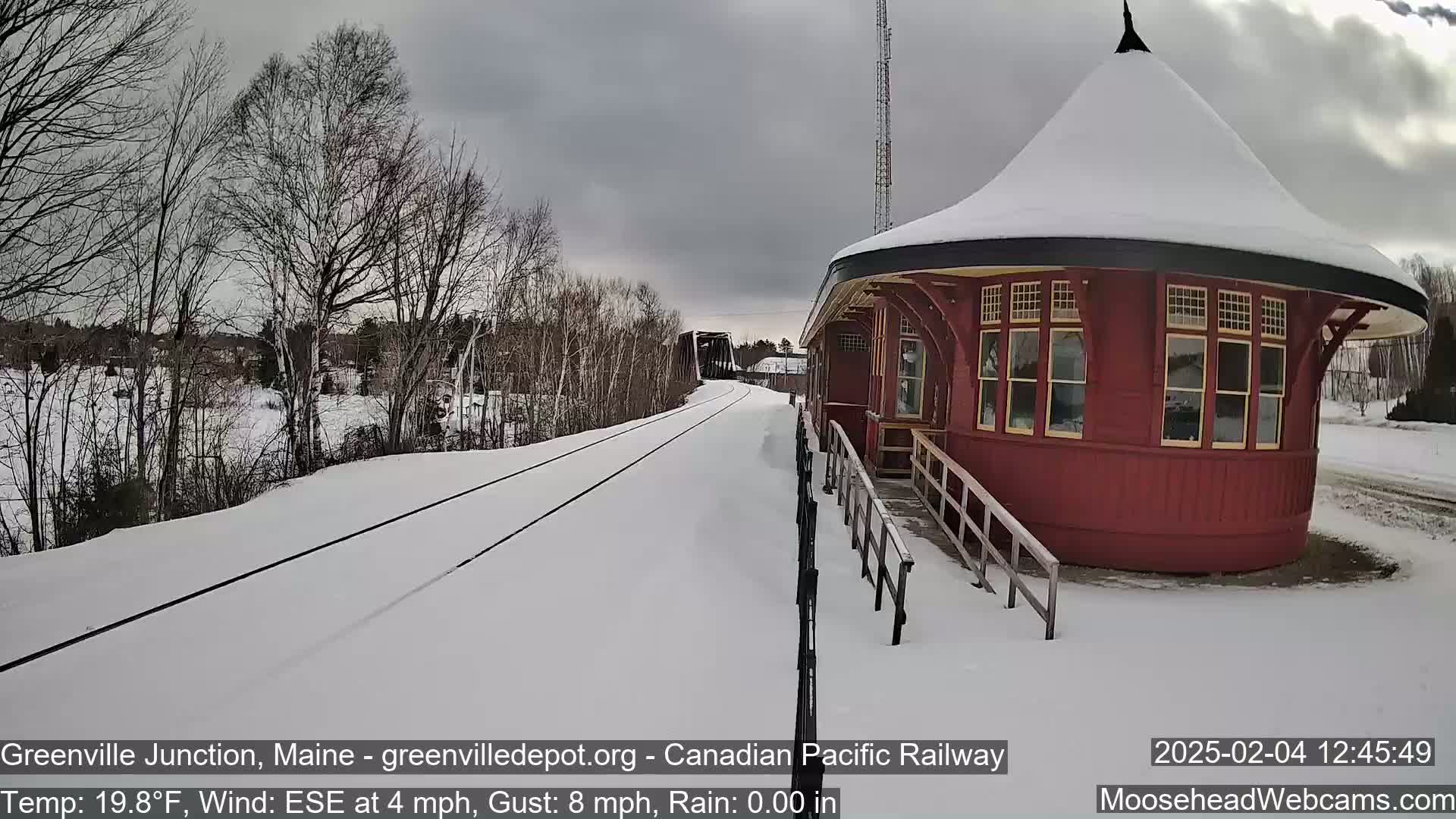 A snow-covered railway track leads to a red, round train station under a cloudy sky.