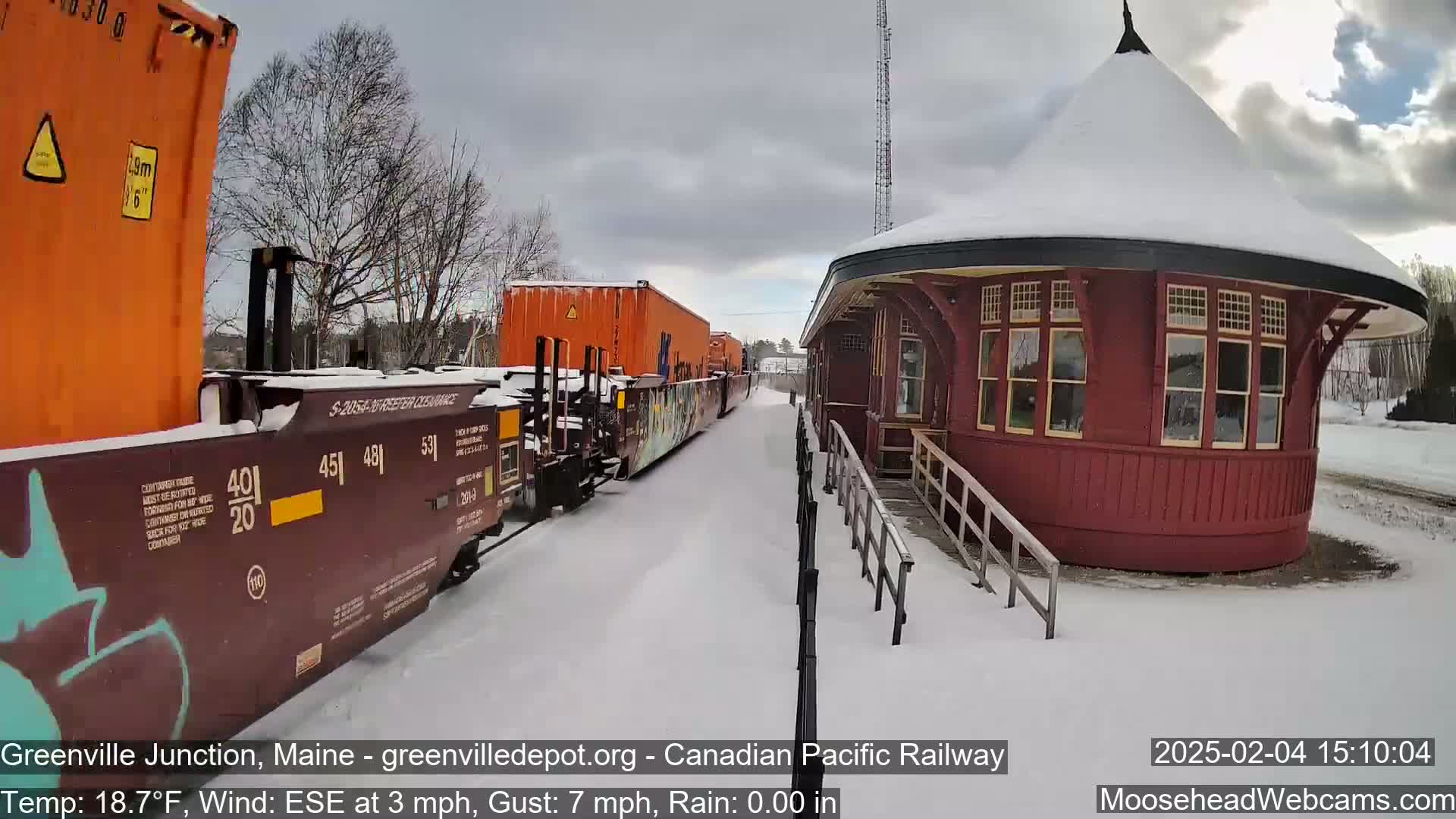 A brown freight train with orange containers pulls into a snow-covered train station under a cloudy sky.