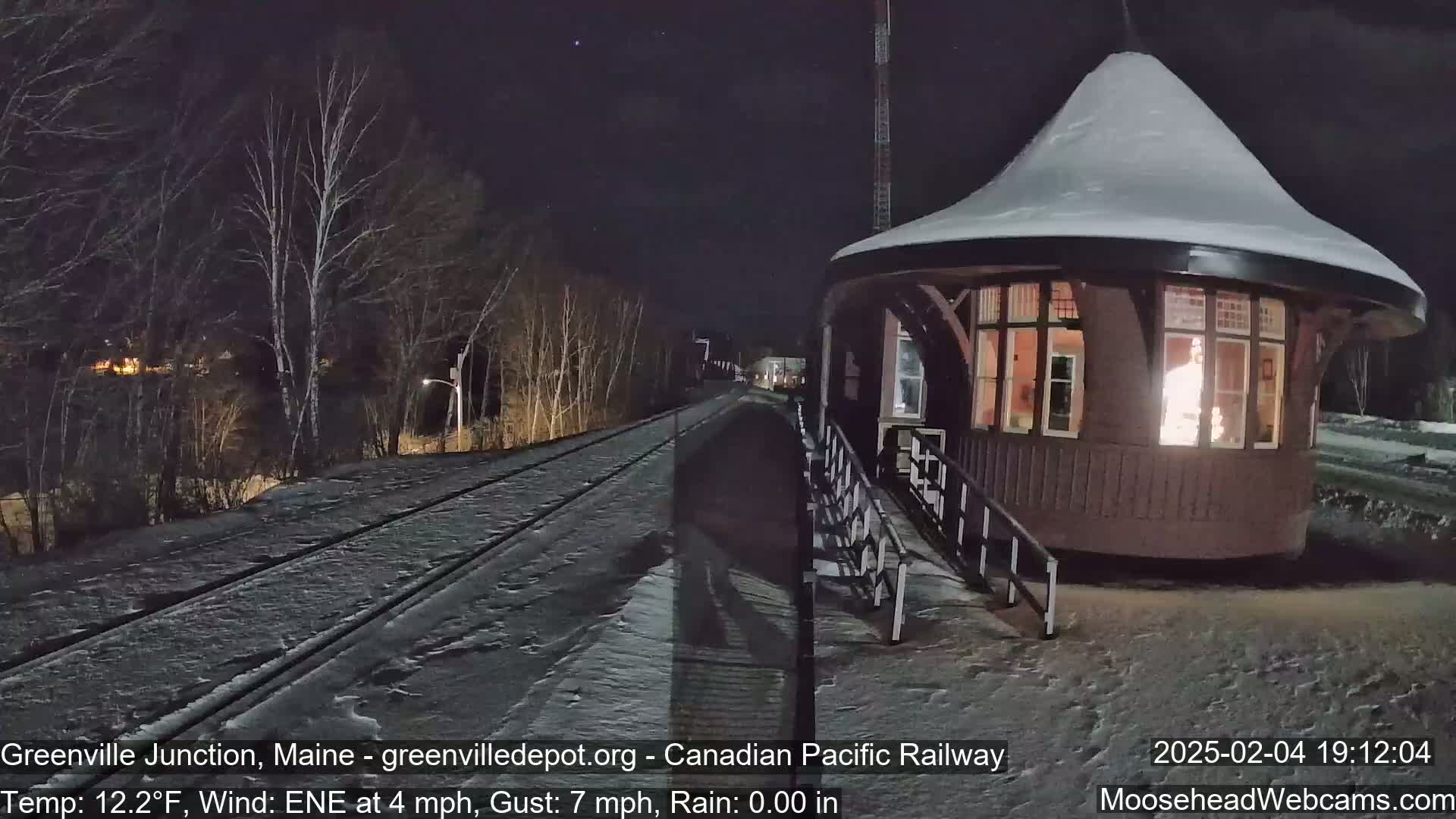 A snow-covered train platform with a lit-up, round building next to a snow-covered railway track at night.