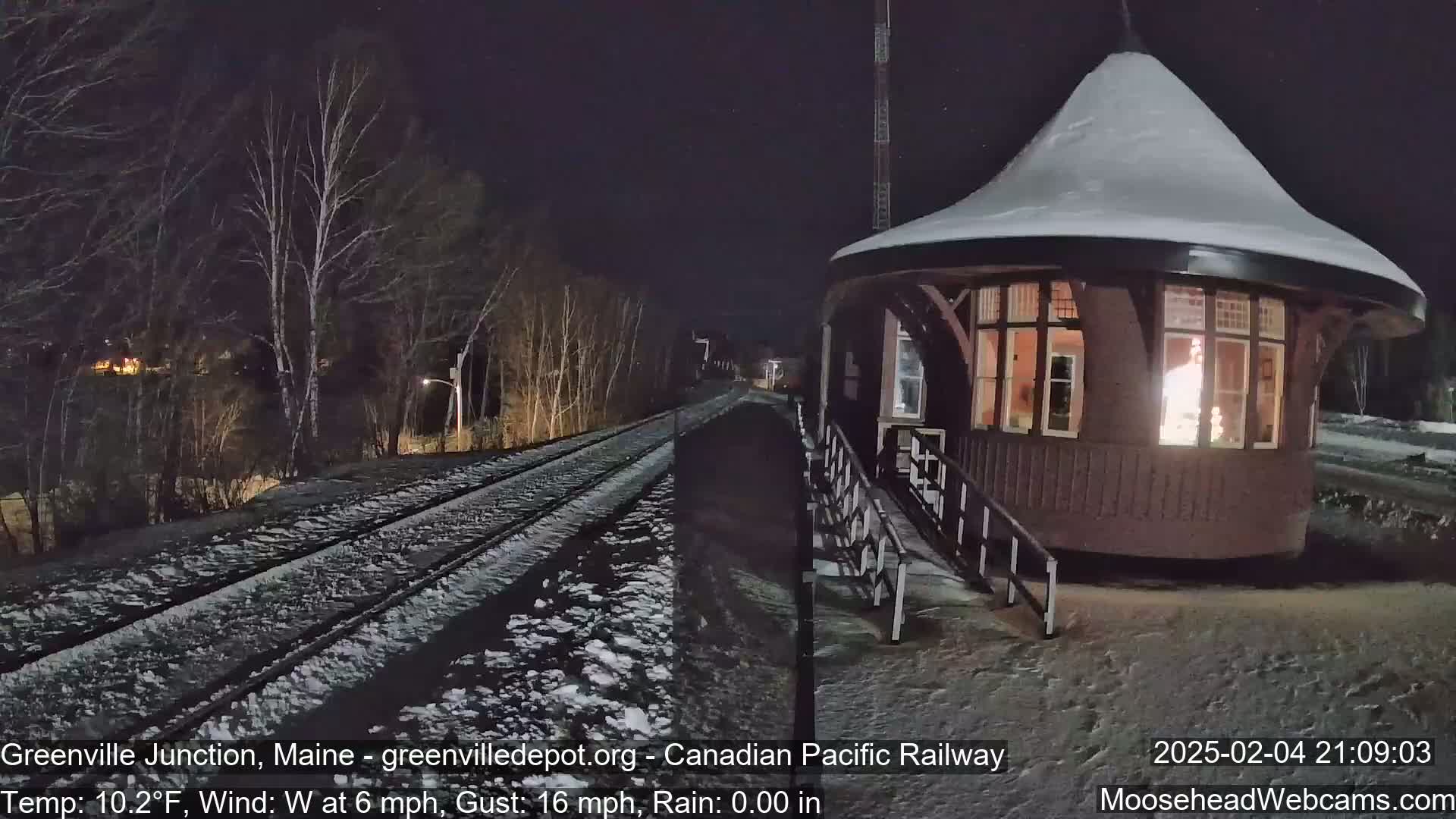 A snow-covered railway track leads to a small, round, illuminated building at night, under a dark sky with a west wind.